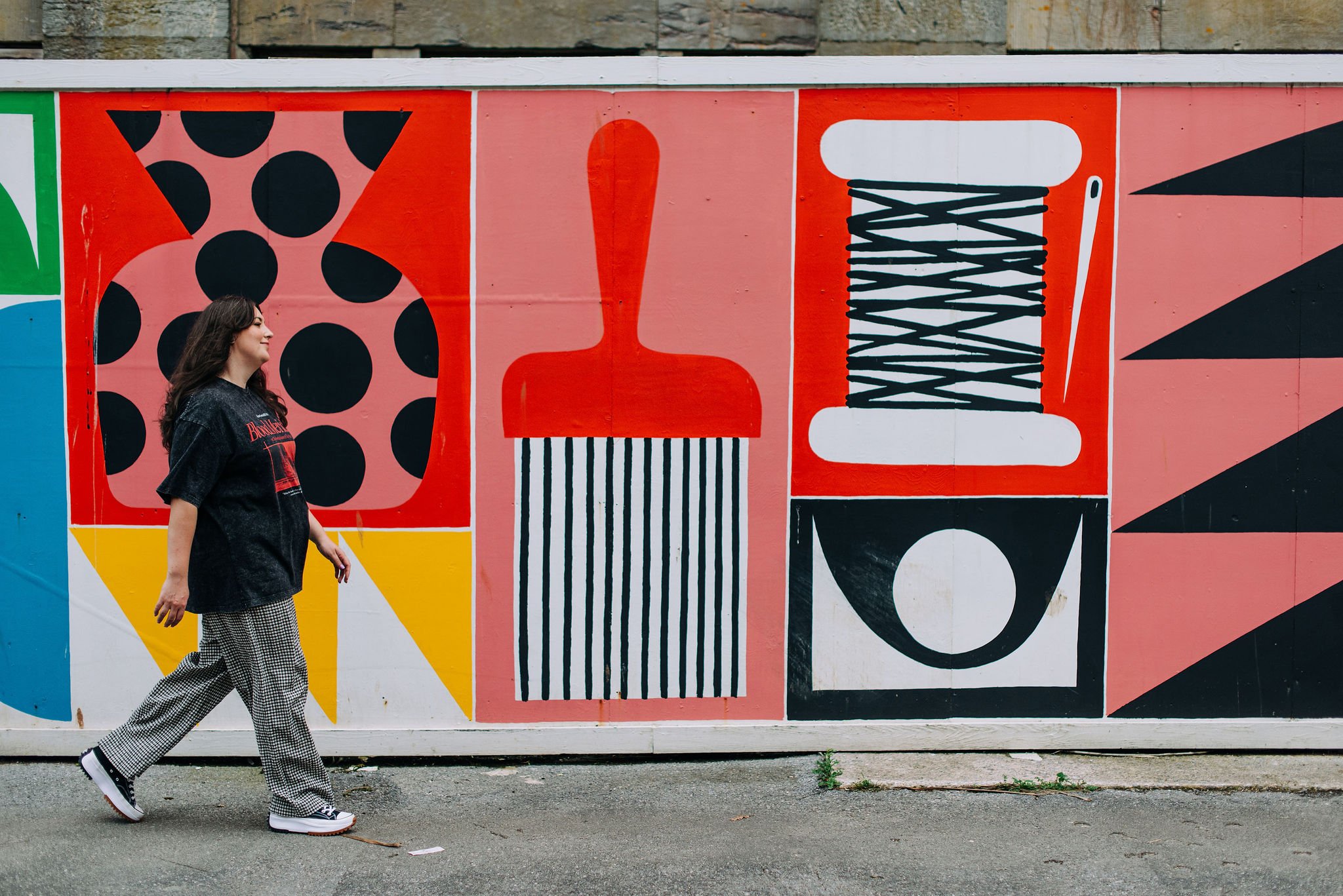 A young woman with long dark hair is walking past a colorful geometric mural on a wall. She is wearing a black T-shirt, checkered wide-leg pants, and black and white sneakers.