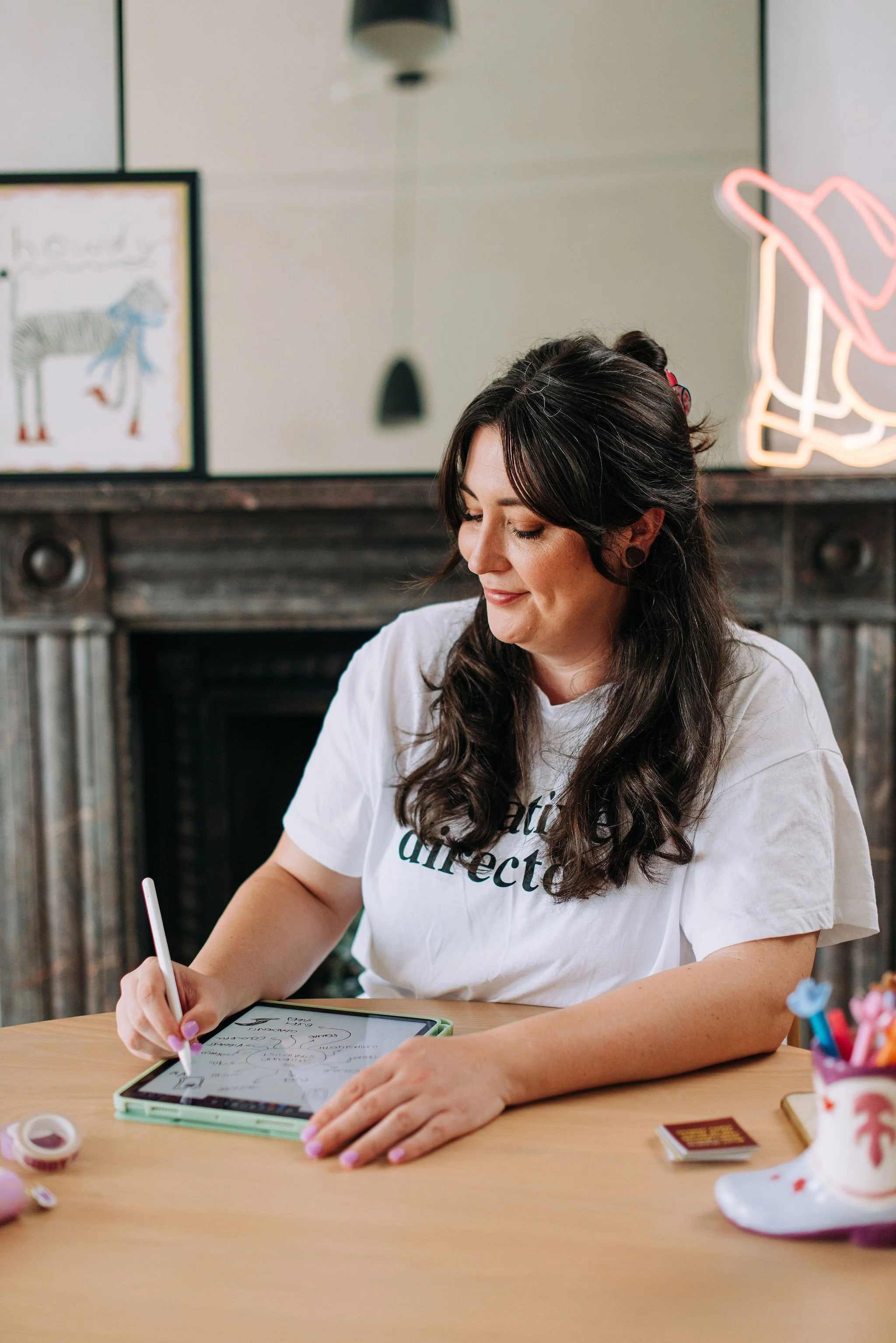 A woman with long dark hair, wearing a white t-shirt, is sitting at a wooden table, writing with a stylus on a digital tablet, smiling softly. There are art supplies and decorations like a pink flamingo figure and a colorful container of pens on the table, with a mirror, framed art, and a wood-paneled wall in the background.