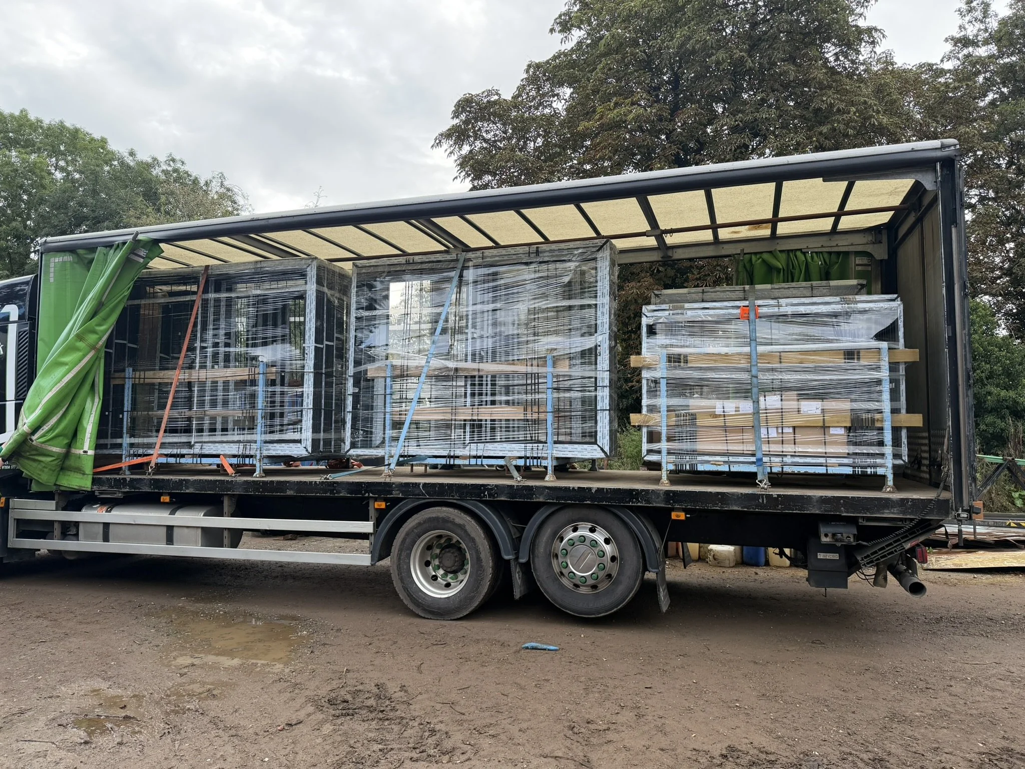 A flatbed truck carrying stacks of wrapped building materials on a dirt ground with trees in the background.