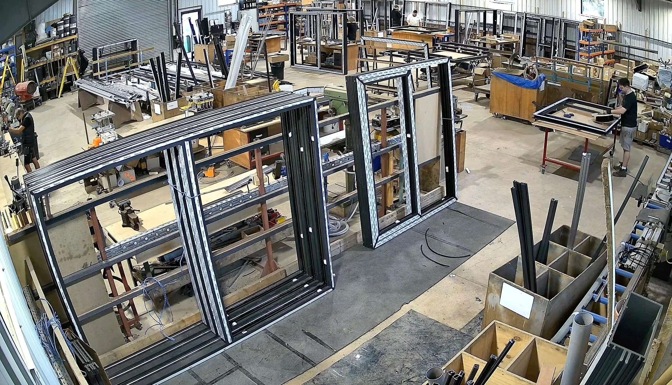 A large workshop with metal frames being assembled, with workers working on different projects, shelves with tools and materials along the walls, and various equipment scattered around.