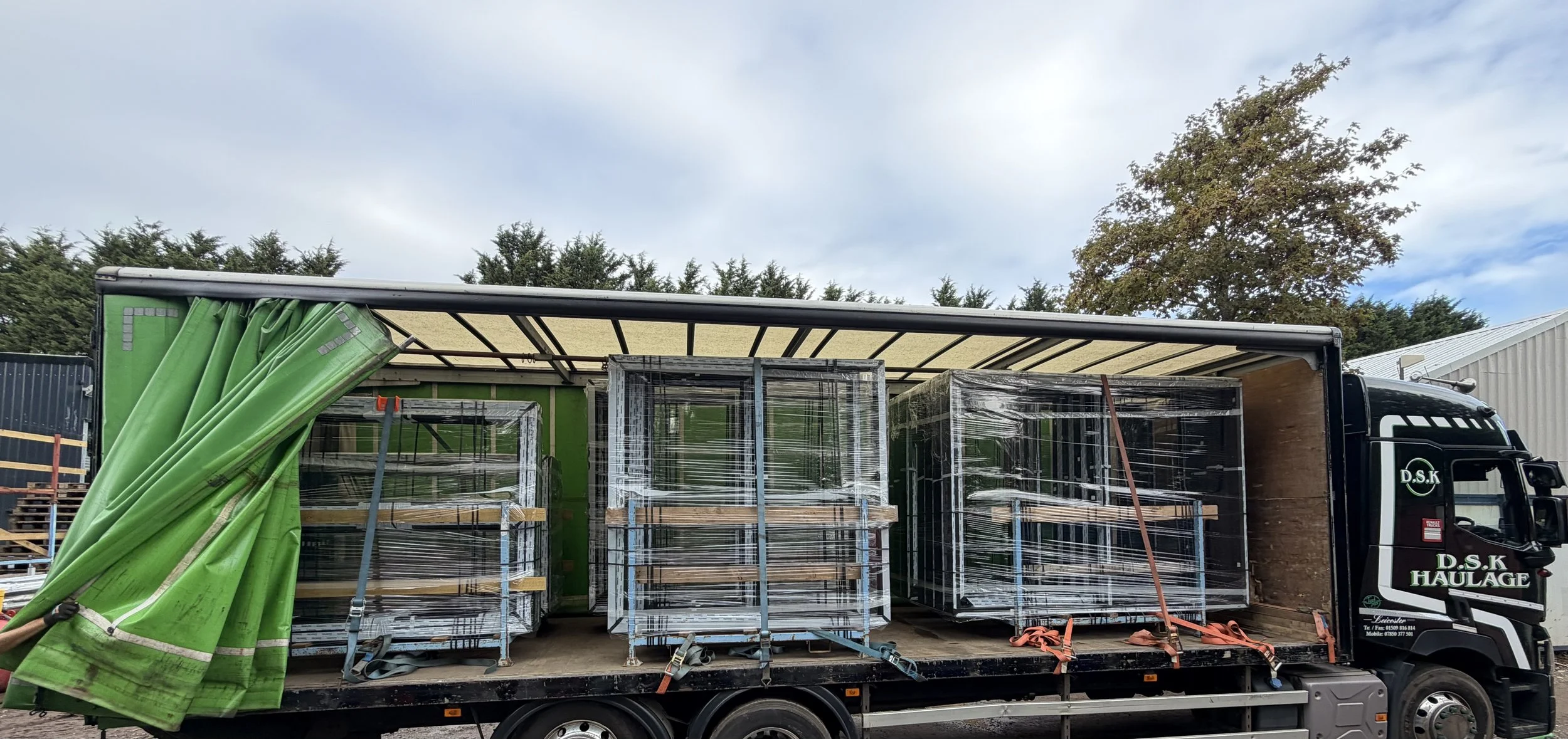 A truck loaded with metal shelving, some wrapped in plastic, parked outdoors with trees and a warehouse in the background.