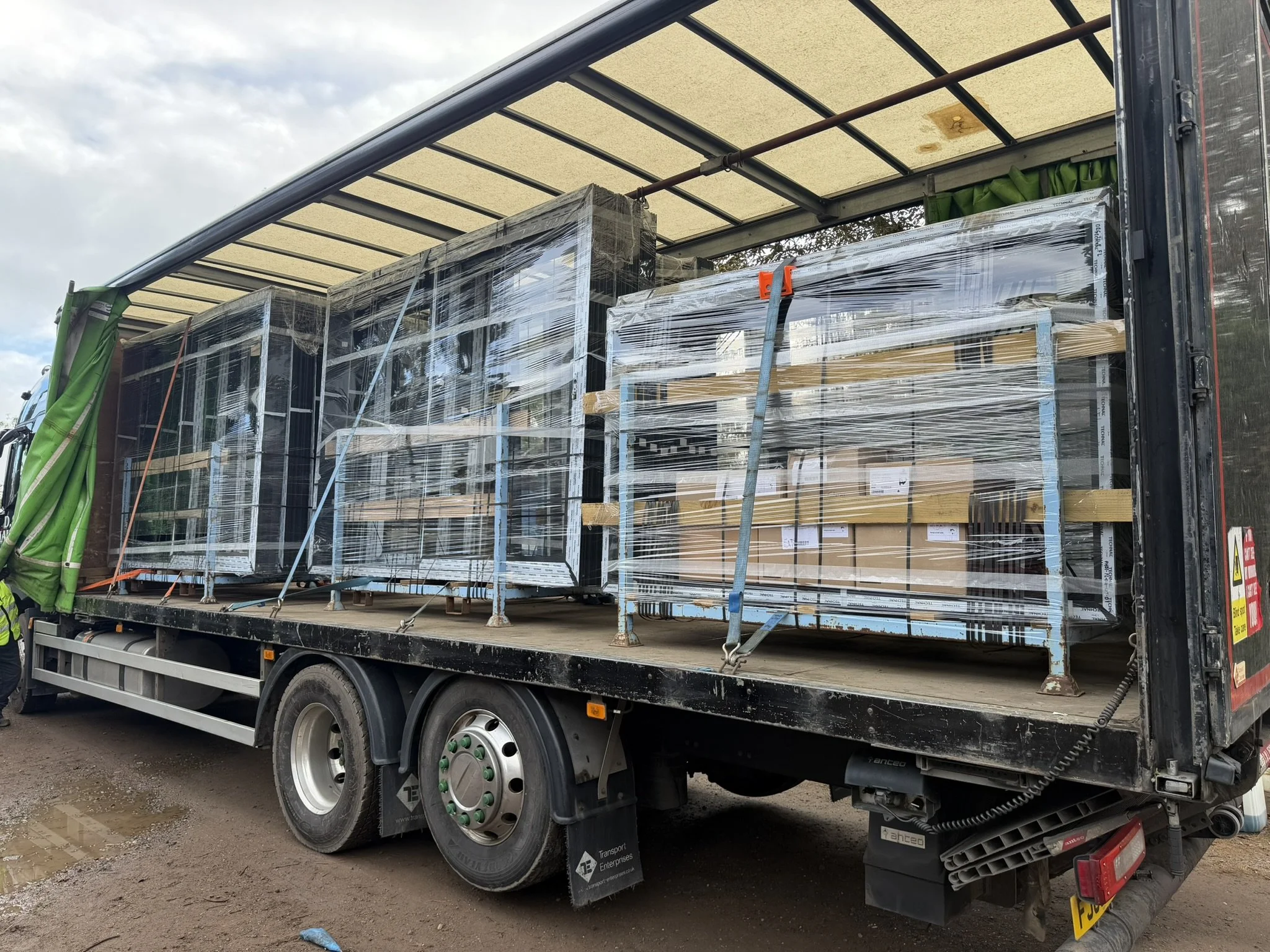 A large truck loaded with wrapped glass windows or doors, parked outdoors on a dirt surface under a cloudy sky.