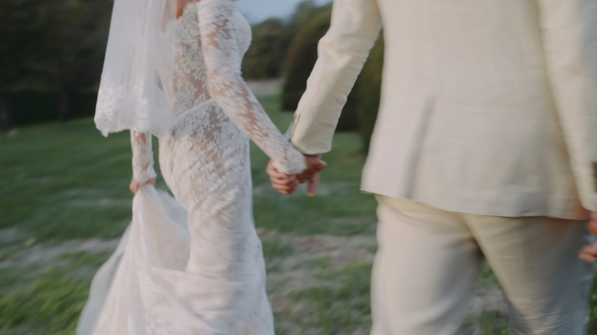 A couple holding hands, walking outside in a grassy area during daylight. The woman is wearing a white lace dress and the man is dressed in a light-colored suit.