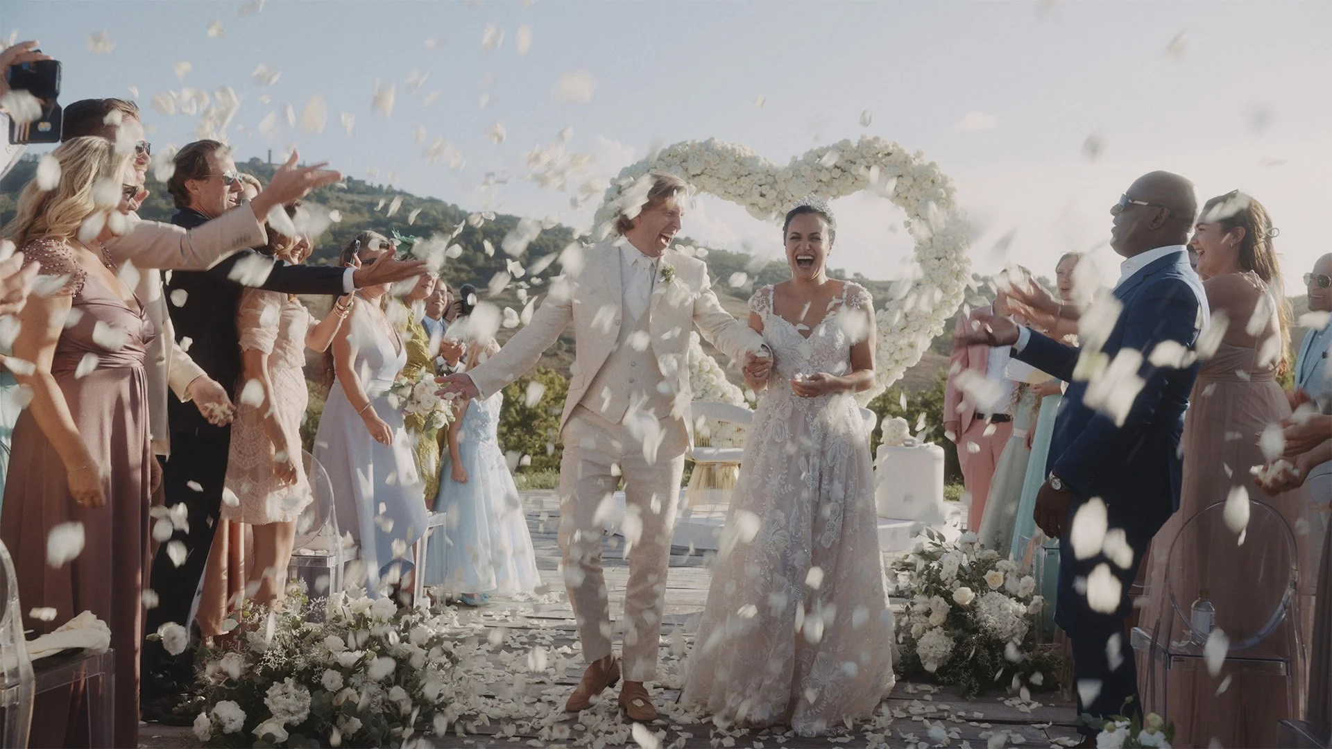 A wedding celebration with a bride and groom walking through guests throwing white flower petals, outdoors on a sunny day, with floral decorations and a heart-shaped floral arch.