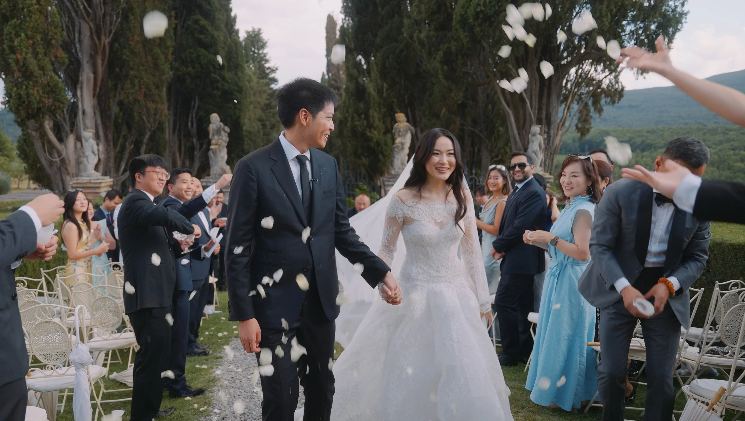 Bride and groom holding hands walking through wedding guests throwing flower petals outdoors, green trees and mountains in background.
