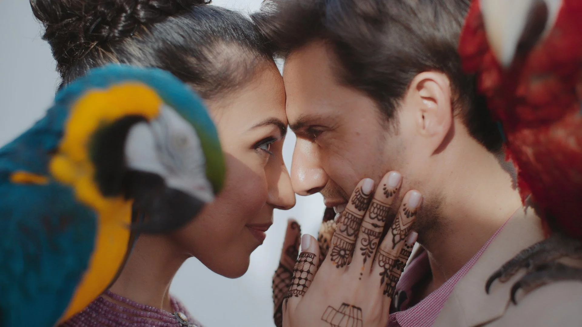 A woman and a man face-to-face with foreheads touching, holding hands with intricate henna designs, a colorful parrot on her shoulder, and people in the background.
