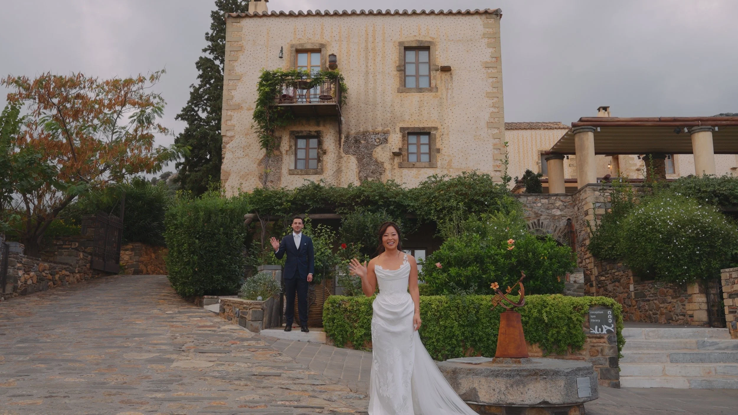Bride in a white wedding dress smiling and waving, standing on a stone pathway in front of a lush green garden, with a man in a dark suit waving behind her, in front of a rustic house with peeling paint and a balcony, during daytime.