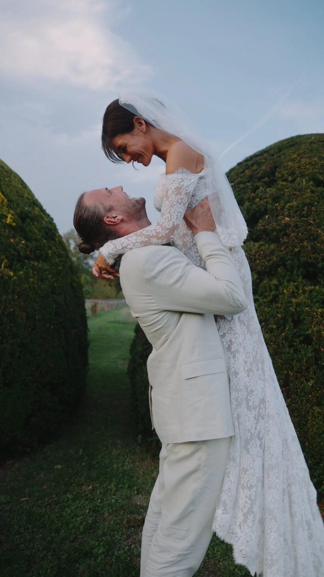 A couple in wedding attire, with the groom lifting the bride in an outdoor garden setting, smiling at each other.