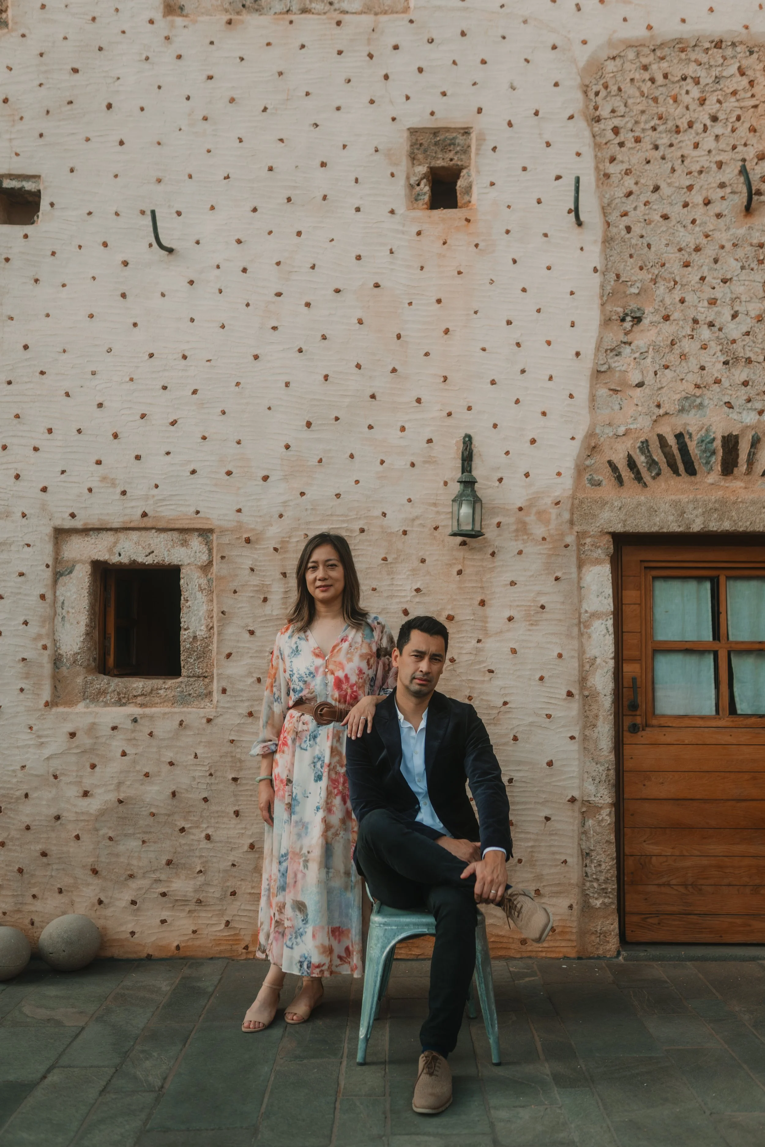 A woman in a long floral dress standing next to a man in a black jacket and pants, sitting on a chair against a textured beige wall with small holes and a window.