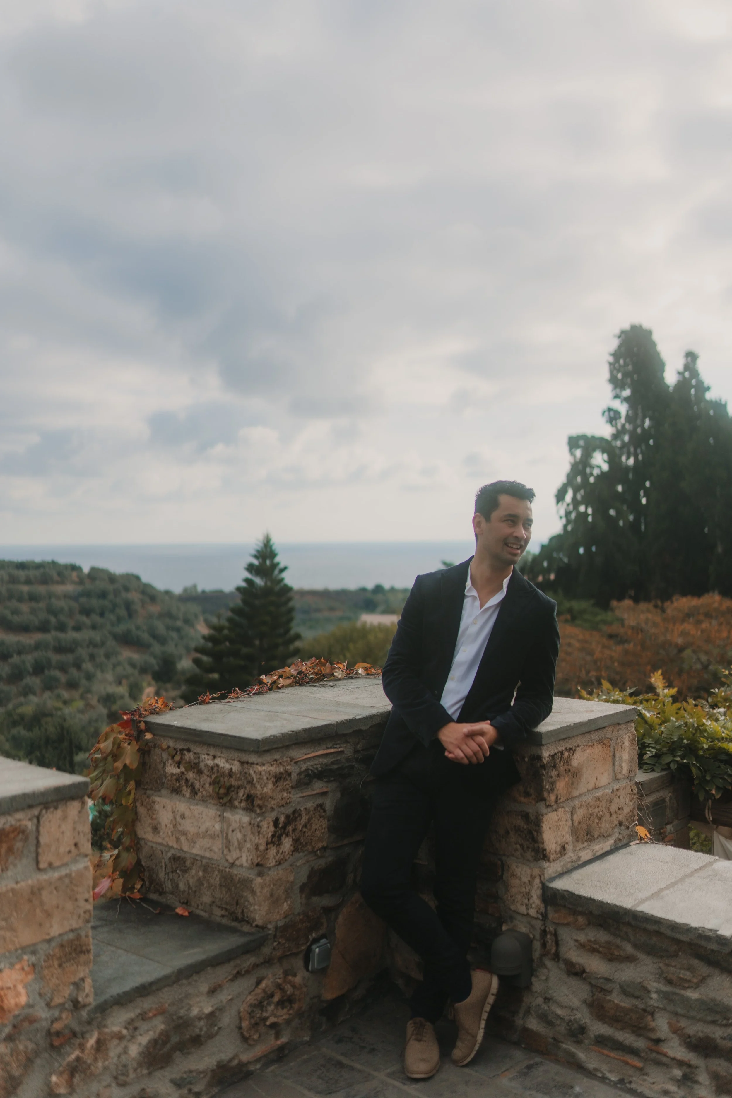 A man in a black suit leaning against a stone wall outdoors, with trees, hills, and the ocean in the background, under a cloudy sky.