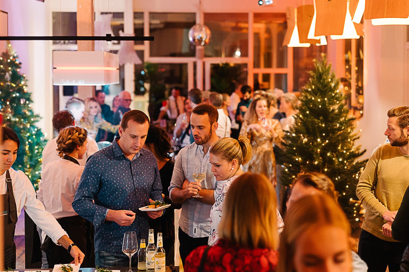 Menschen feiern Weihnachten bei einem festlichen Abendessen, Schneeflocken und Weihnachtsbäume im Hintergrund, warmes Licht und festliche Dekorationen.