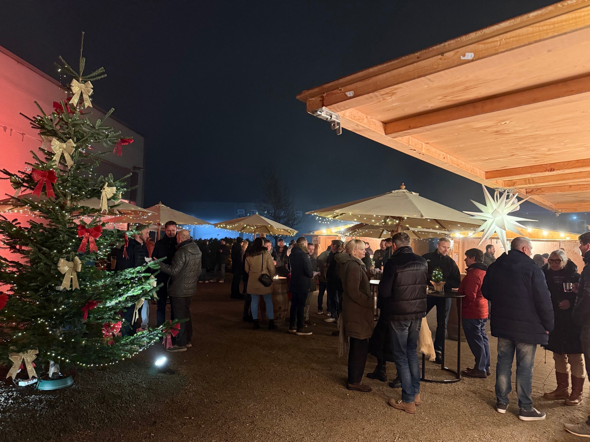 Menschen versammeln sich bei einer Weihnachtsfeier im Freien bei Nacht, mit Weihnachtsbaum, Lichterketten und Sterndeko.