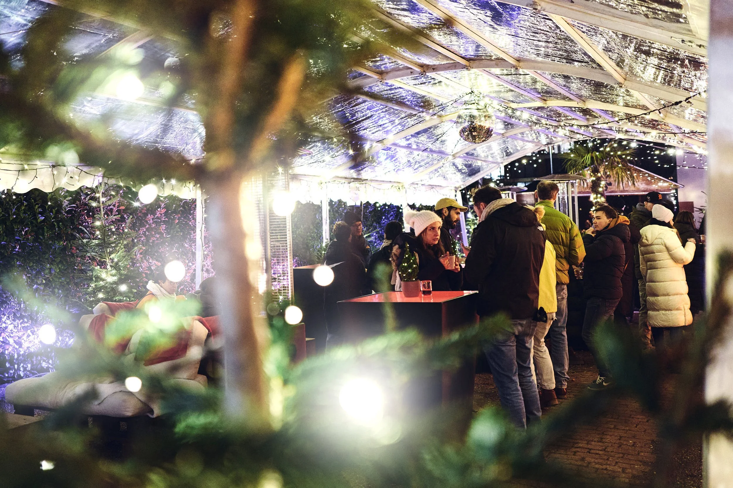 Menschen feiern bei einer festlich dekorierten Weihnachtsfeier unter einem transparenten Dach mit Lichterketten und einer Weihnachtsbeleuchtung.