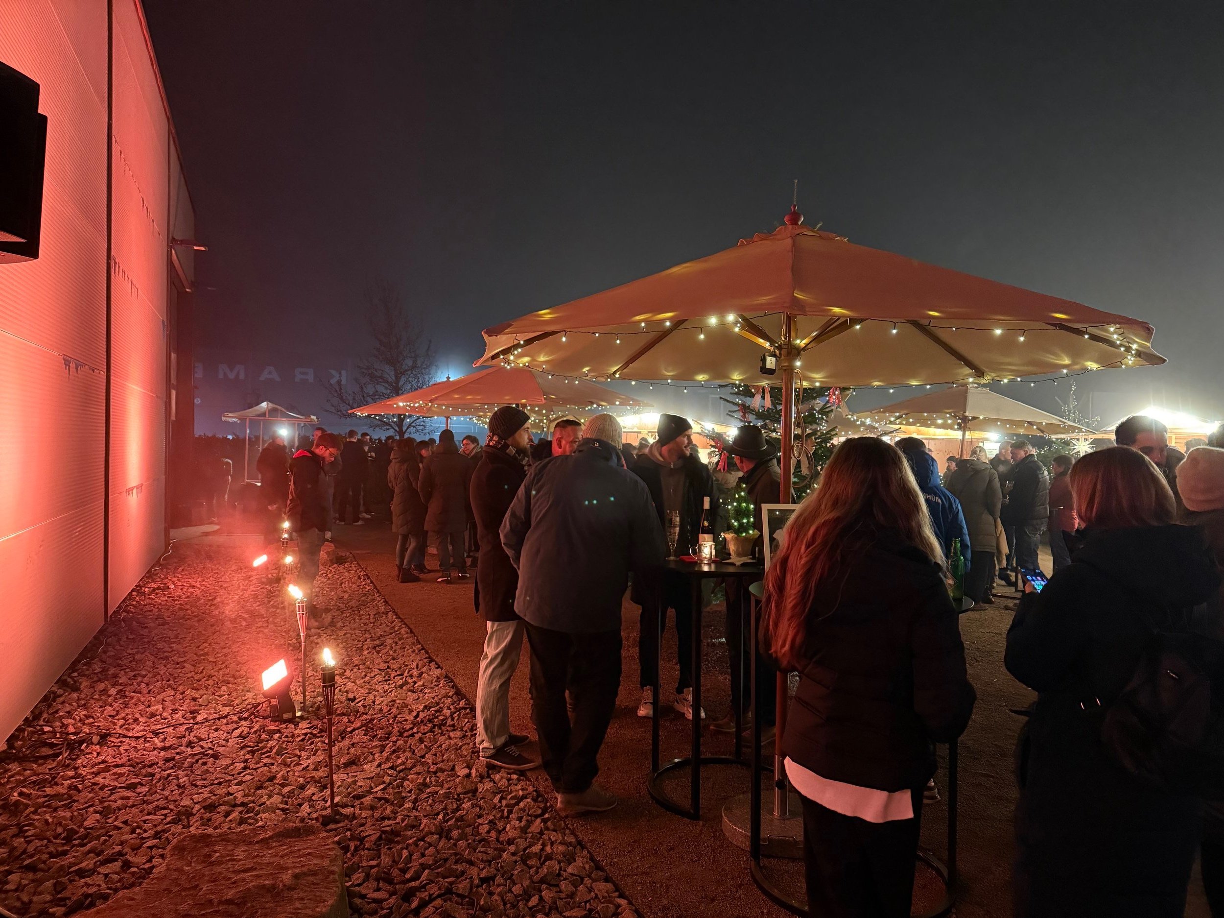Menschen versammeln sich bei einer festlichen Veranstaltung im Freien bei Nacht, unter großen Schirmen mit Lichterketten, mit Weihnachtsbaum und Lichter im Hintergrund, umgeben von kleinen Feuerstellen, auf einem befestigten Außenbereich.
