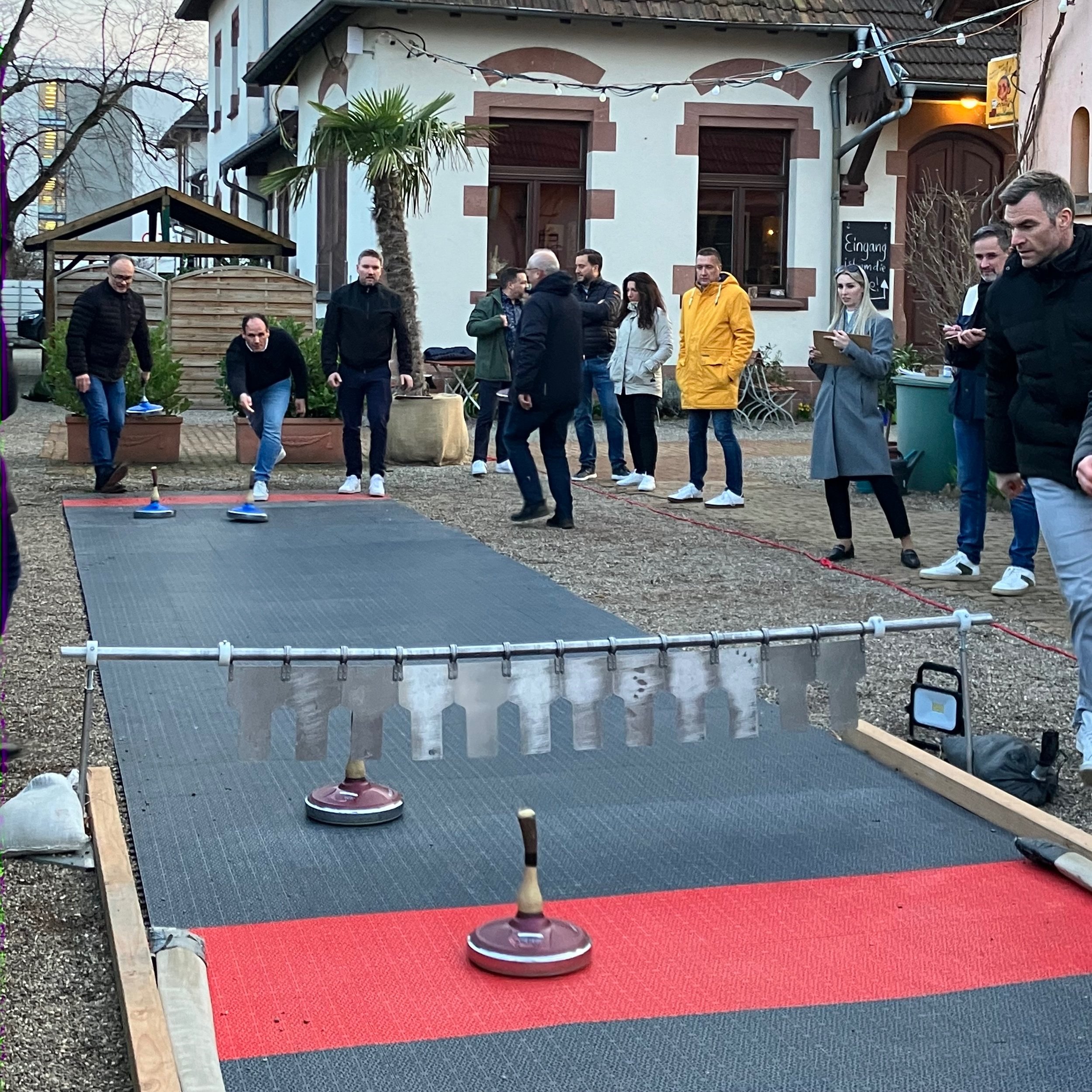 Mehrere Menschen spielen eine Curling-ähnliche Sportart auf einer improvisierten Bahn im Freien, umgeben von Gebäuden und gelegentlichen Zuschauern.