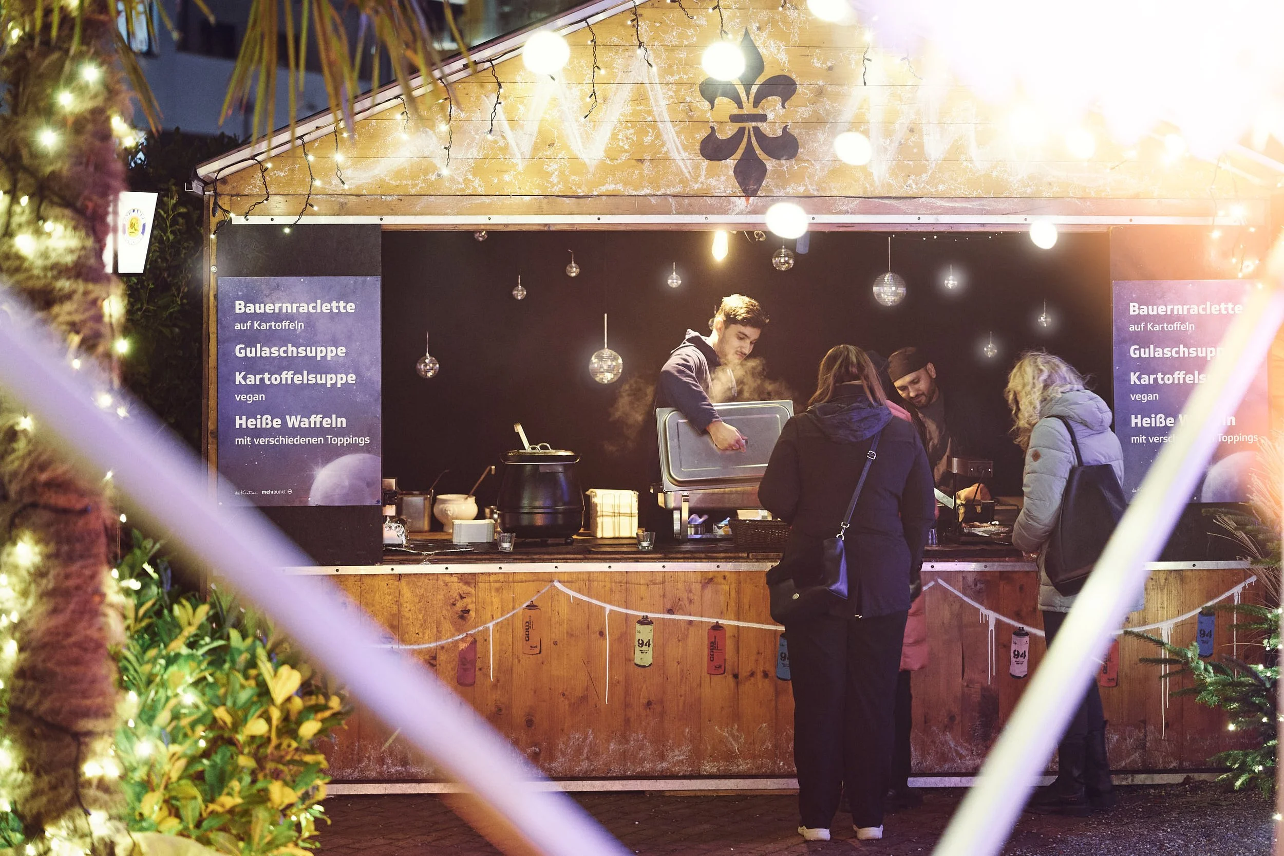 Eine festlich beleuchtete Bude auf einem Weihnachtsmarkt, an der mehrere Menschen Speisen bestellen, darunter eine Ofen- oder Frittenbude mit Speisekarten, die Bakso, Gulaschsuppe, Kartoffelsuppe und heiße Waffeln anbieten.