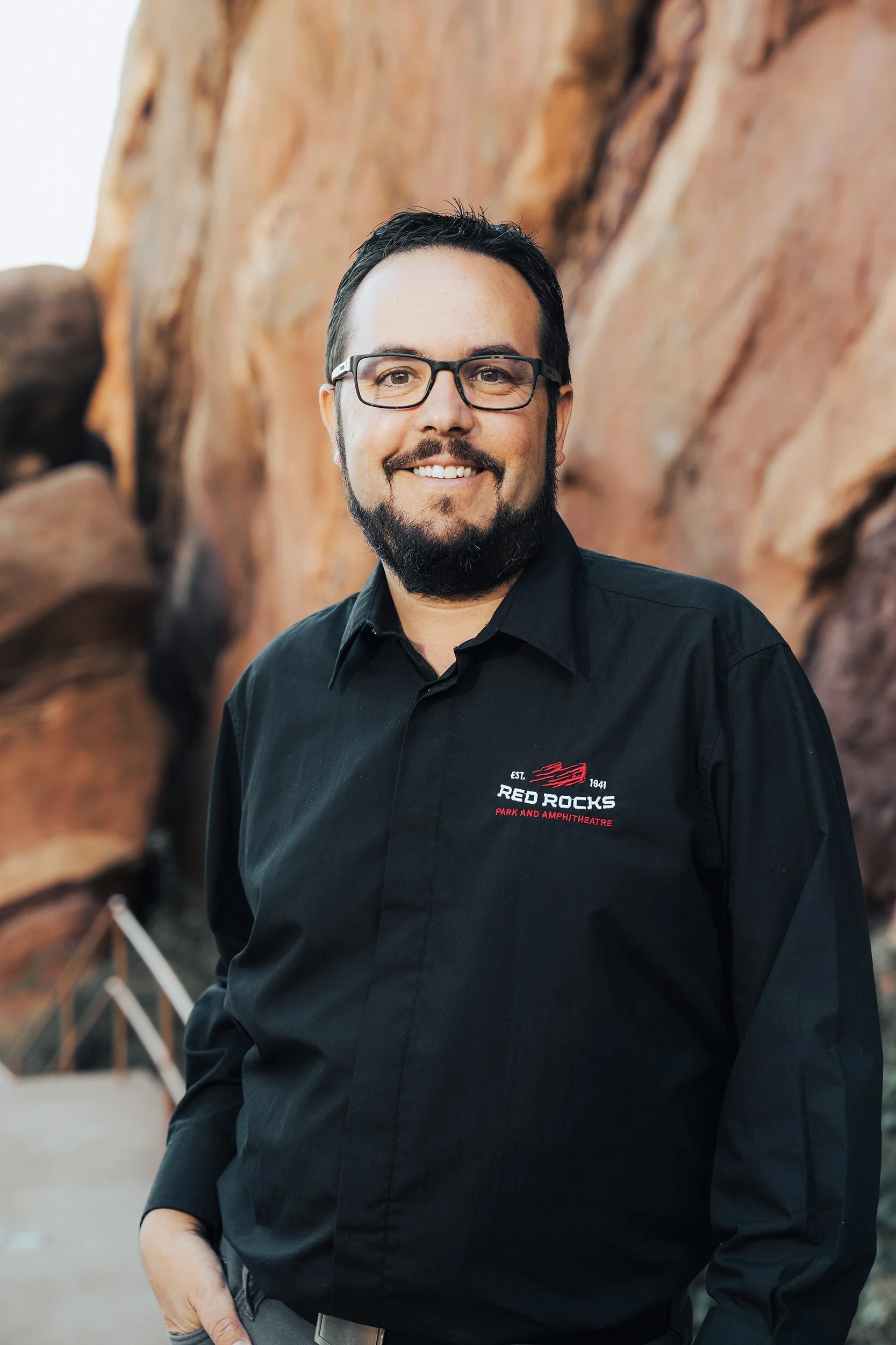 A man with glasses, a beard, and dark hair smiling, wearing a black shirt with a logo that says "Red Rocks Park and Amphitheatre" in front of a rocky background.