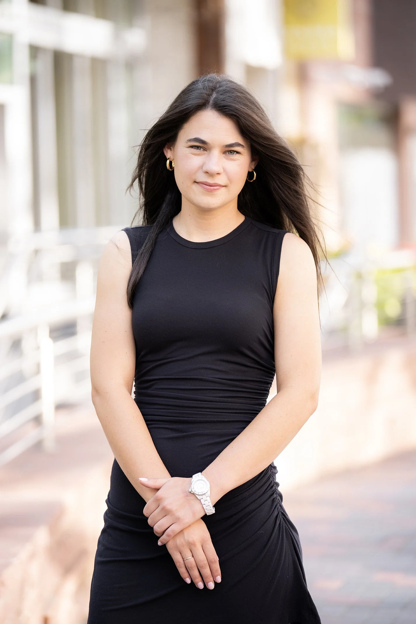 A young woman with long dark hair wearing a sleeveless black dress and gold hoop earrings, standing outdoors on a city street, smiling gently at the camera.