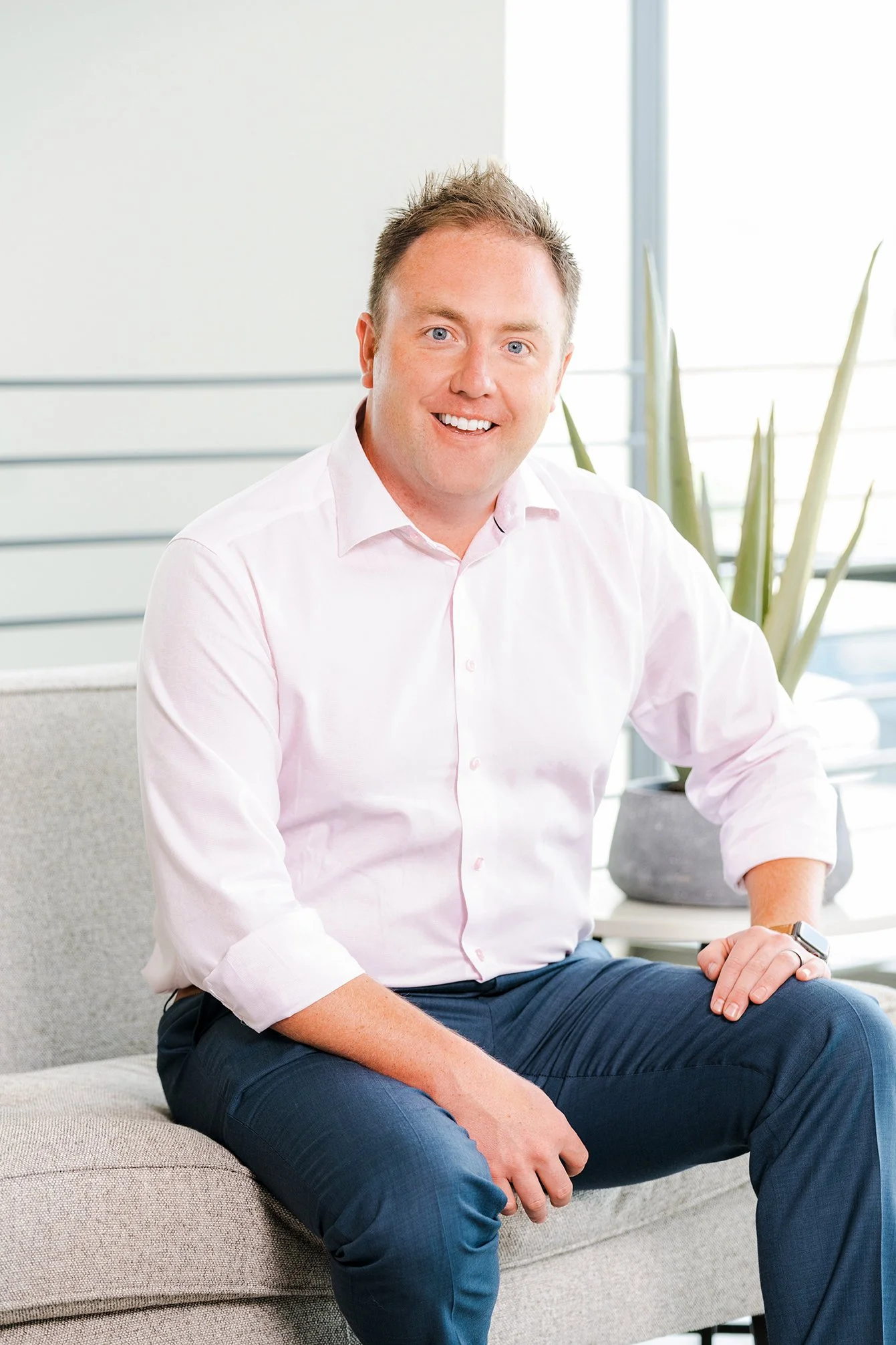 A man with short, light brown hair wearing a light pink button-up shirt and dark blue pants sitting on a beige couch in a modern office space with large windows and a potted plant in the background.