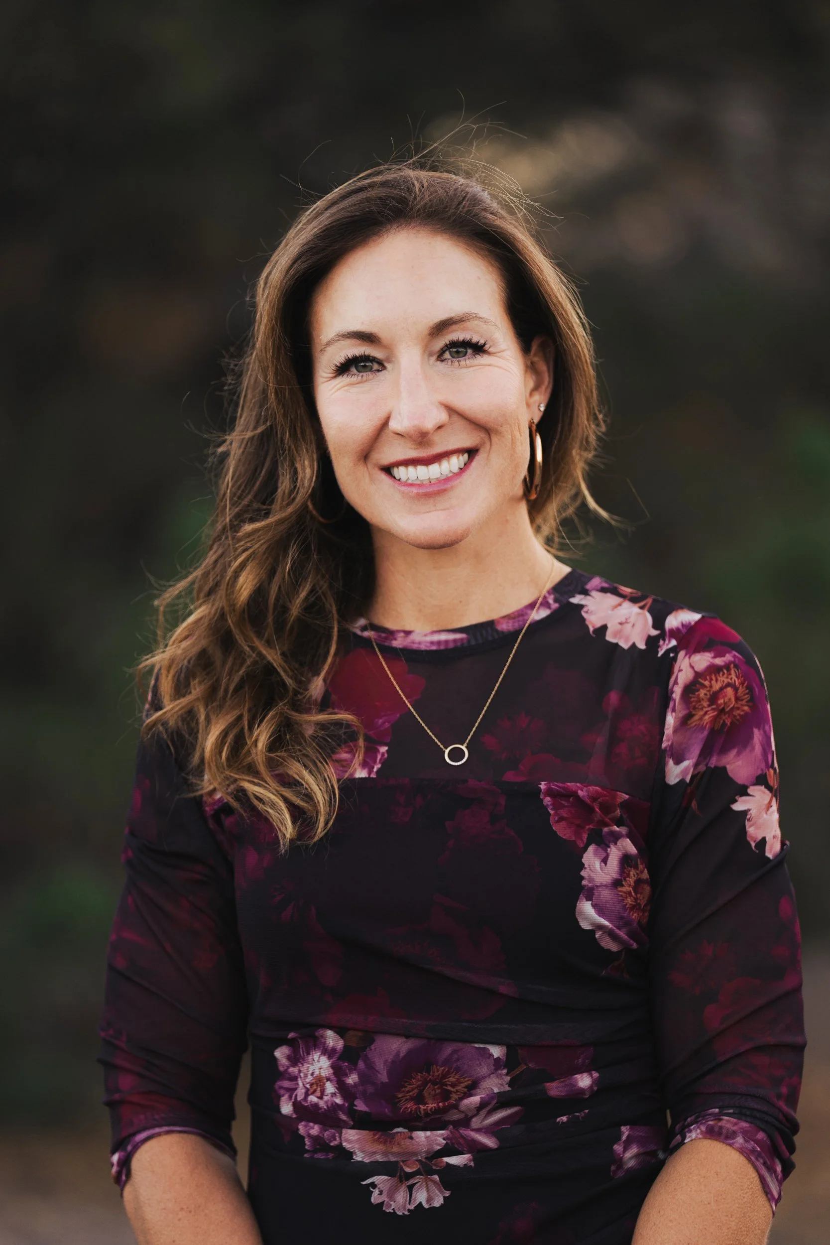 A woman with long wavy brown hair and fair skin, smiling, wearing gold hoop earrings, a gold necklace with a circular pendant, and a dark floral top, outdoors.