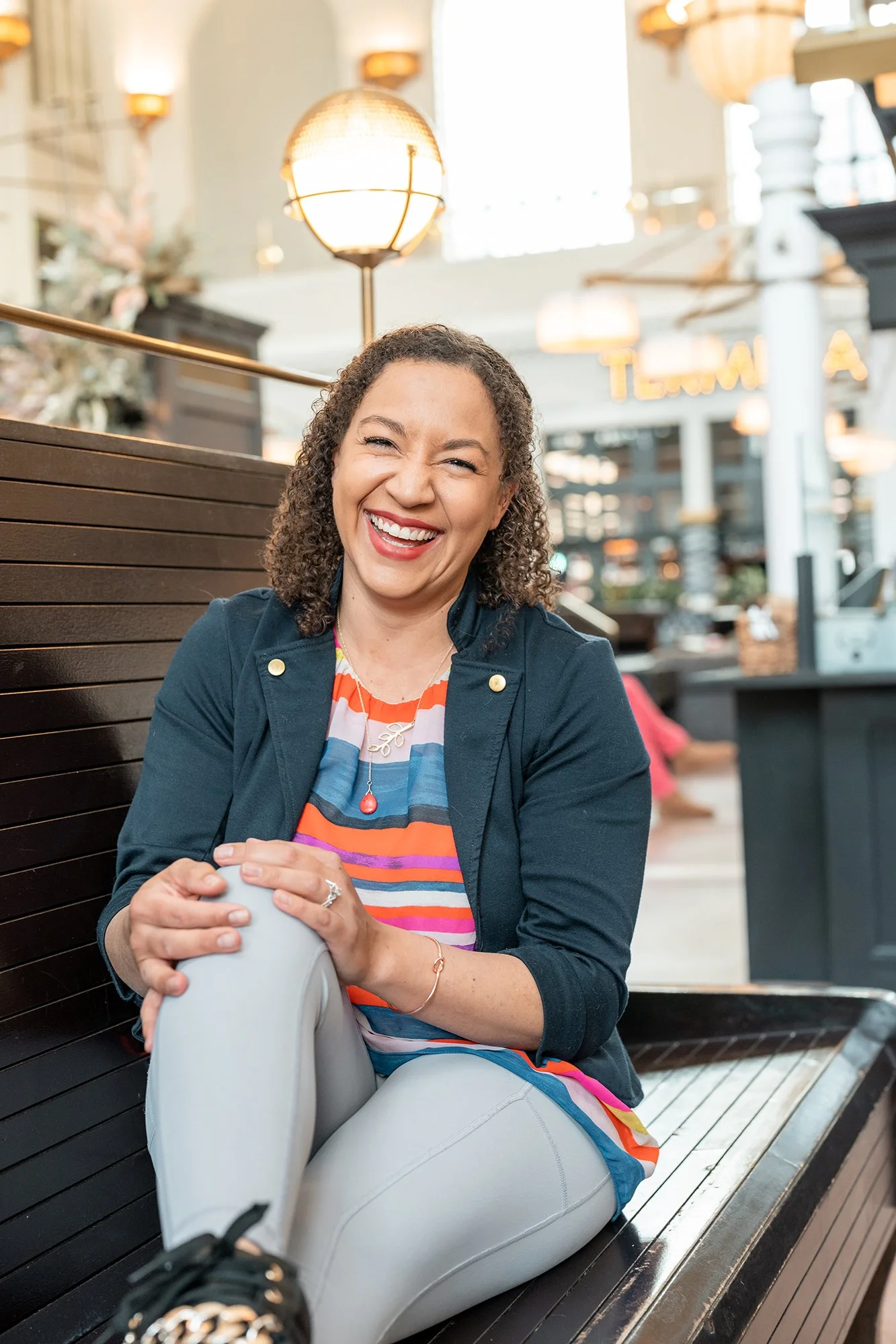 A woman with curly hair smiling, sitting on a bench in a bright indoor setting with hanging lights and a floral arrangement in the background.