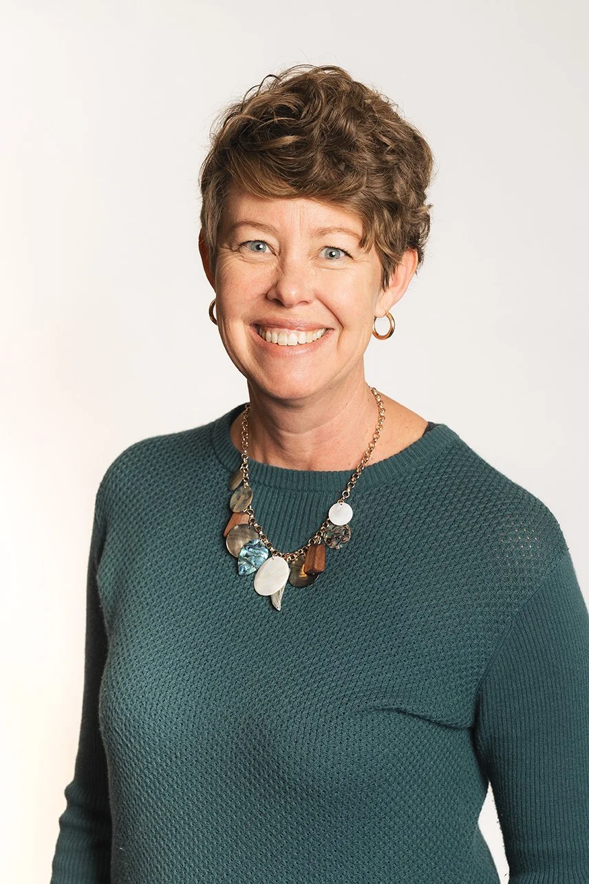 Portrait of a smiling woman with short brown hair, wearing hoop earrings, a chunky necklace with large shell and stone pieces, and a teal knit sweater, standing against a plain white background.