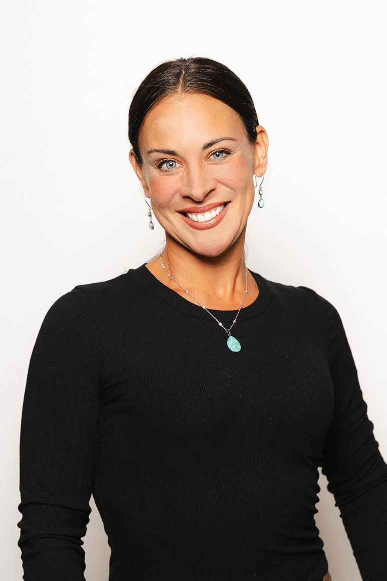 A woman with dark hair tied back, wearing a black long-sleeve top and turquoise jewelry, smiling against a white background.