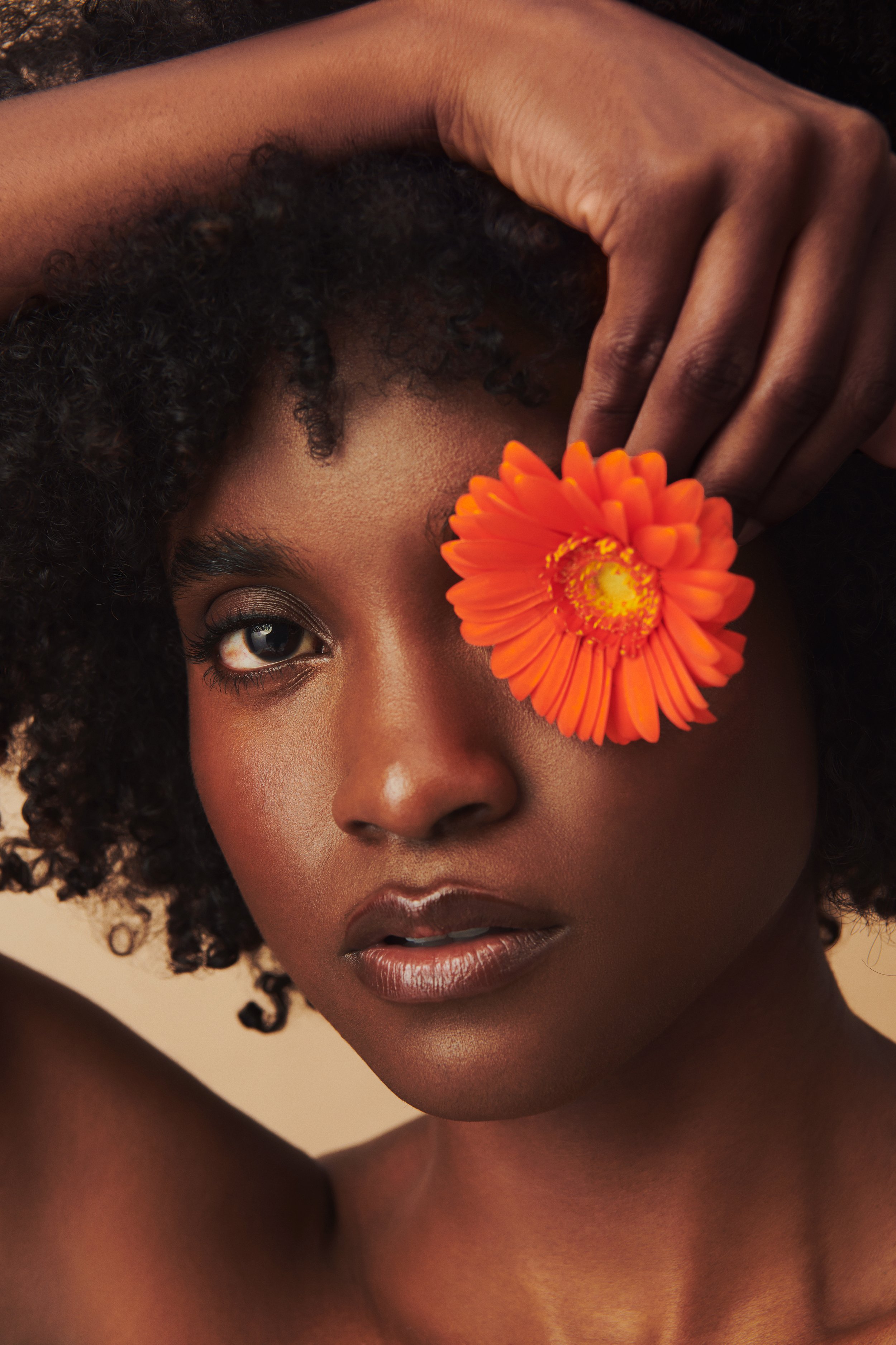 Commercial product photography featuring a model holding an orange flower to her eye shot by QUEST Production Co., London fashion photographers. The model is set against a beige background.