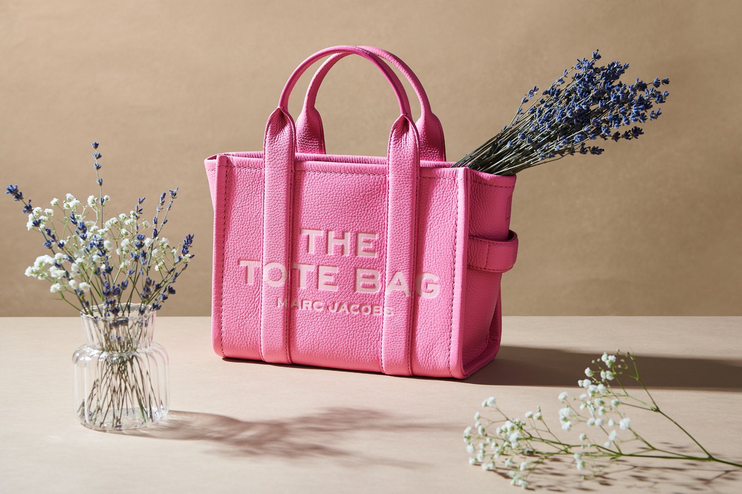 Commercial product photography of Marc Jacobs' pink tote bag shot by QUEST Production Co., London fashion photographers. The bag is displayed with delicate flowers against a beige background.