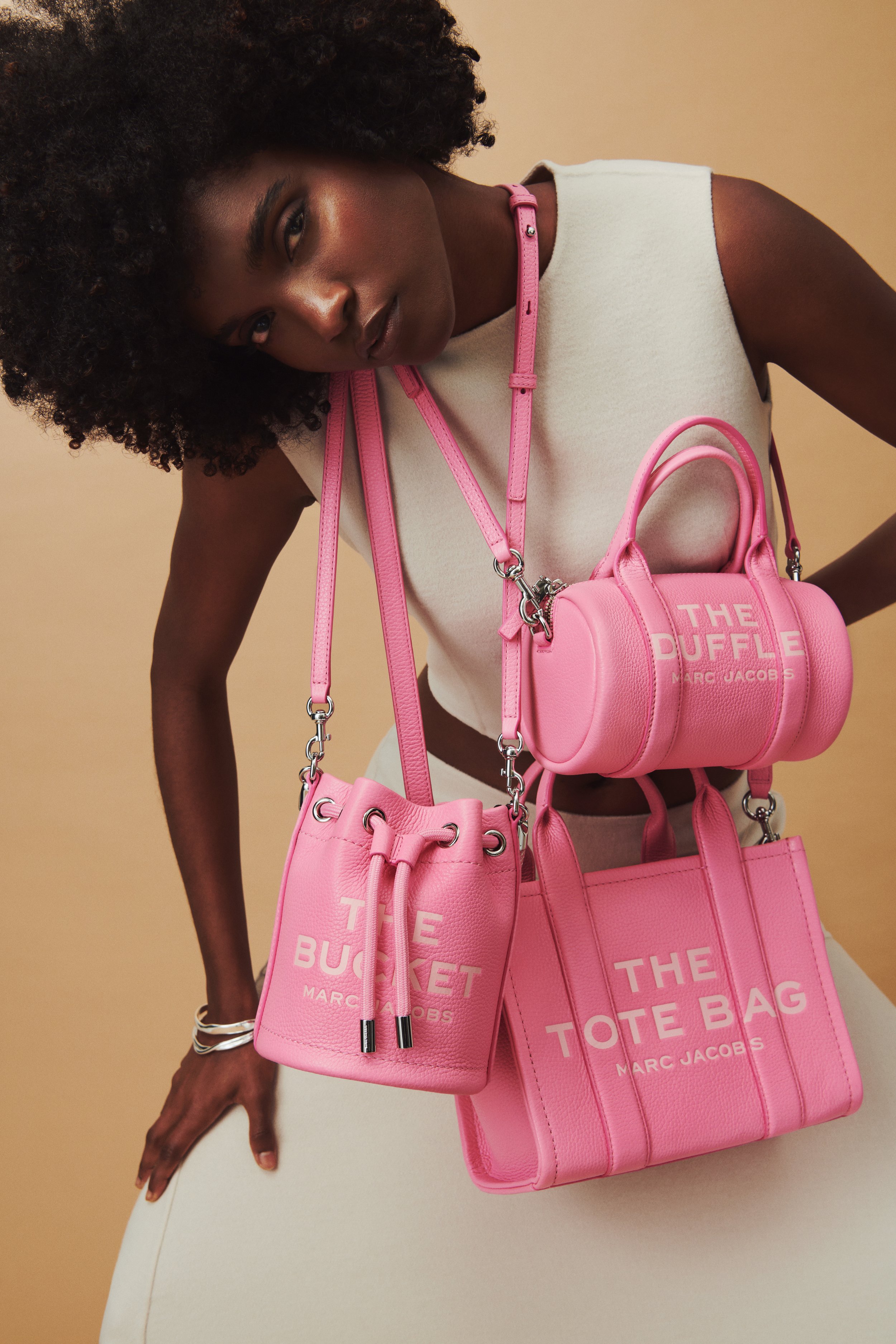 Commercial fashion photography of Marc Jacobs' pink tote, bucket, and duffle bags shot by QUEST Production Co., London fashion photographers. Model in a beige outfit holding the bags against a beige background.