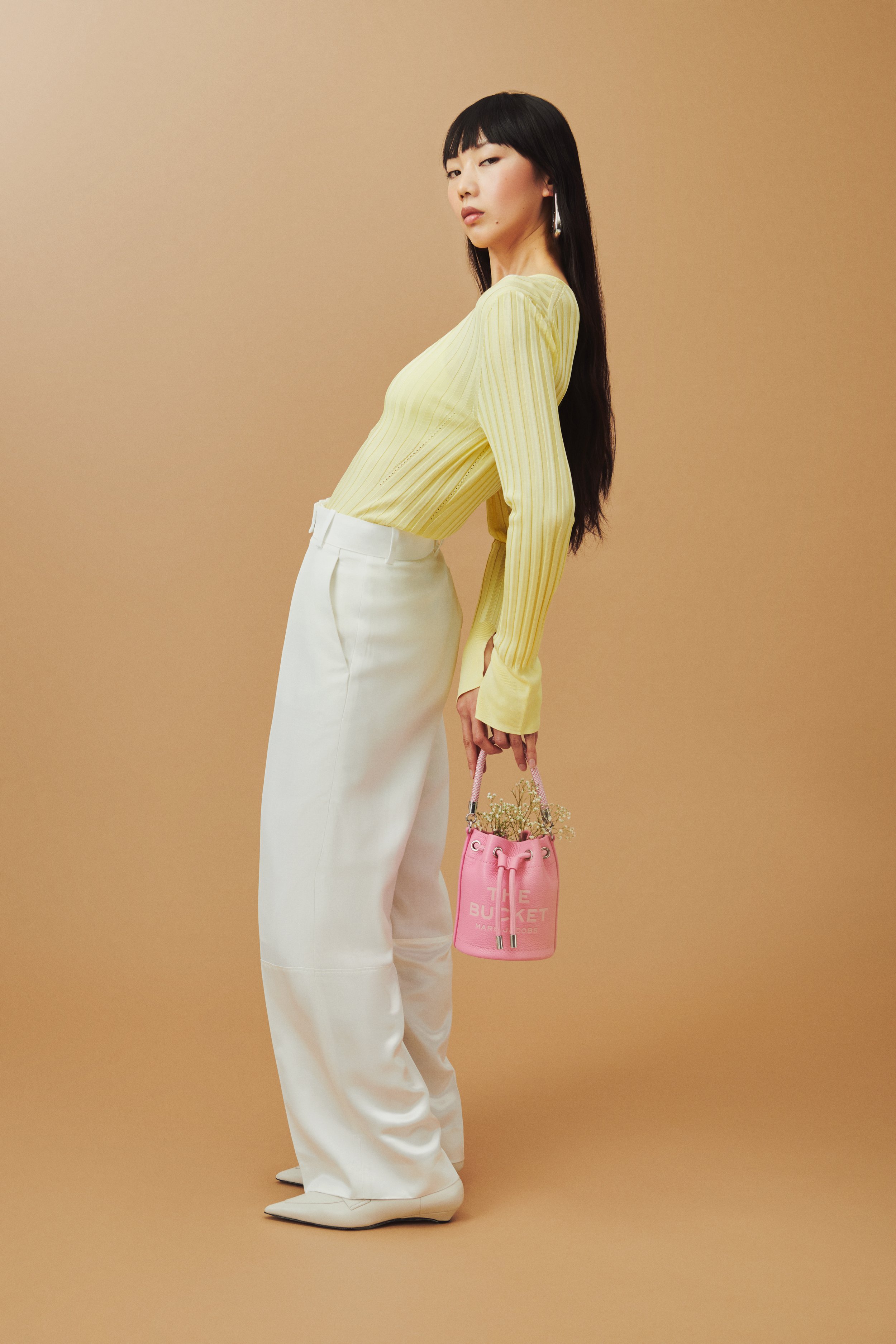 Commercial fashion photography of Marc Jacobs' pink bucket bag shot by QUEST Production Co., London fashion photographers. Model in a yellow top and white pants holding the bag against a beige background.