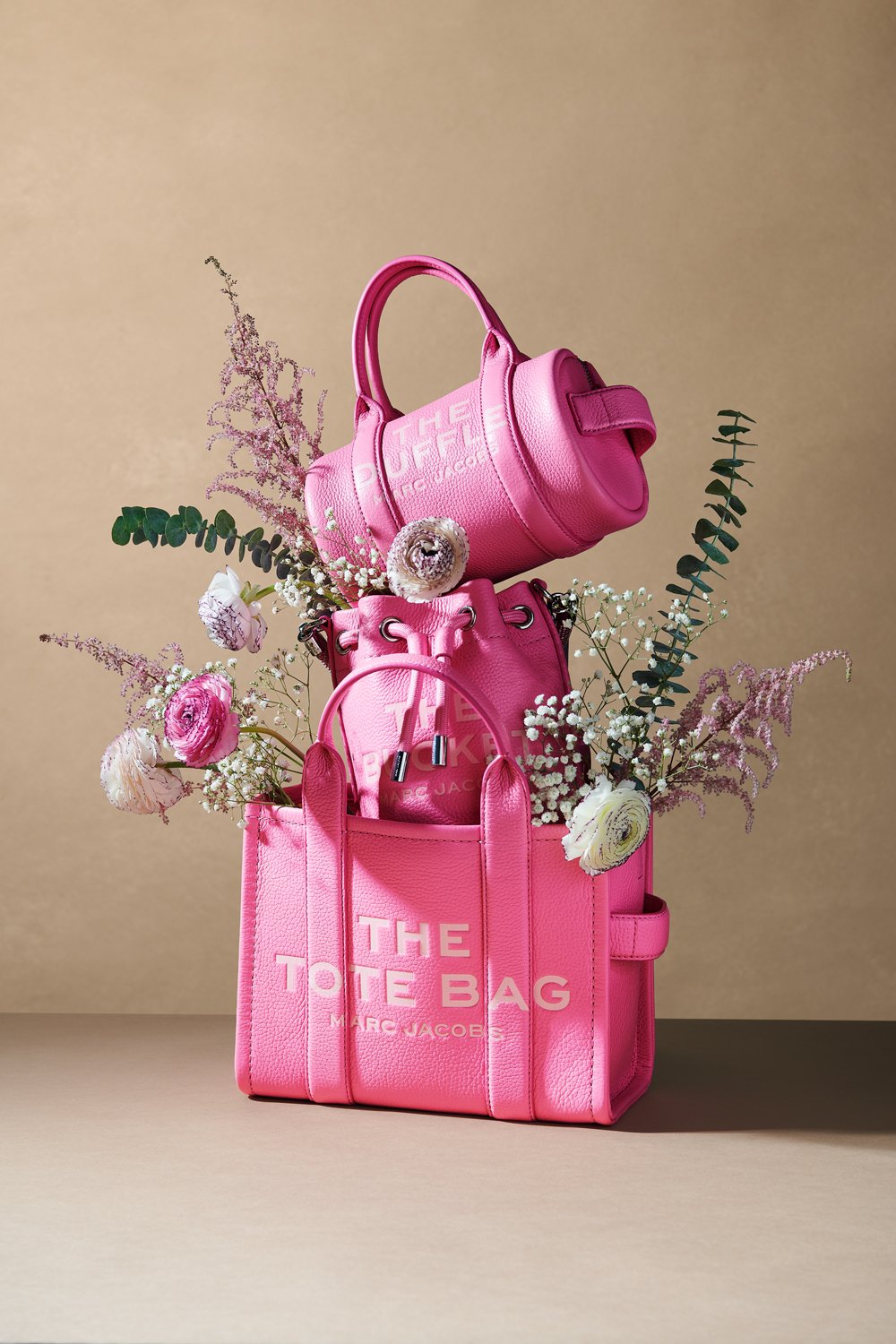Commercial product photography of Marc Jacobs' pink tote, bucket, and duffle bags adorned with flowers shot by QUEST Production Co., London fashion photographers. The bags are displayed against a beige background.