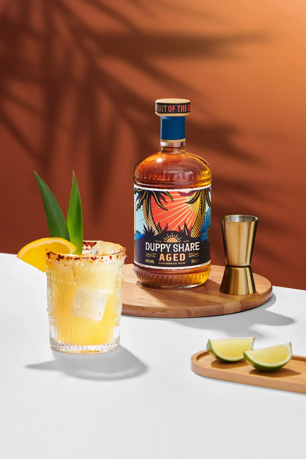 Commercial product photography of Duppy Share Aged Caribbean Rum shot by QUEST Production Co., London fashion photographers. The bottle is displayed with a cocktail and garnishes against a warm orange background.