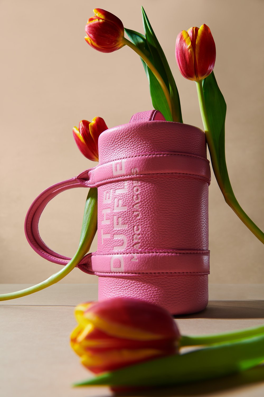 Commercial product photography of Marc Jacobs' pink duffle bag adorned with tulips shot by QUEST Production Co., London fashion photographers. The bag is displayed against a beige background.
