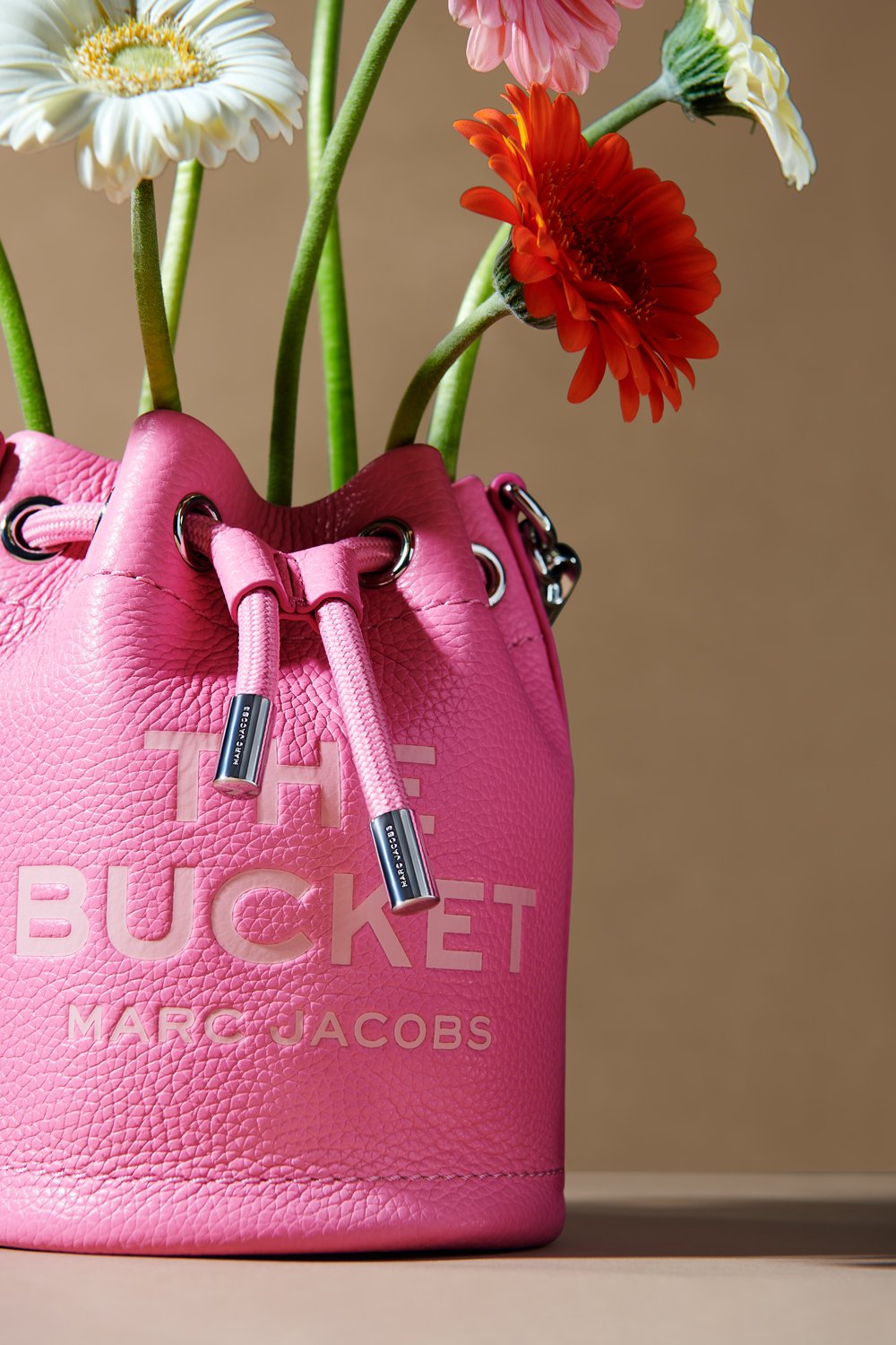 Commercial product photography of Marc Jacobs' pink bucket bag adorned with colorful flowers shot by QUEST Production Co., London fashion photographers. The bag is displayed against a beige background.