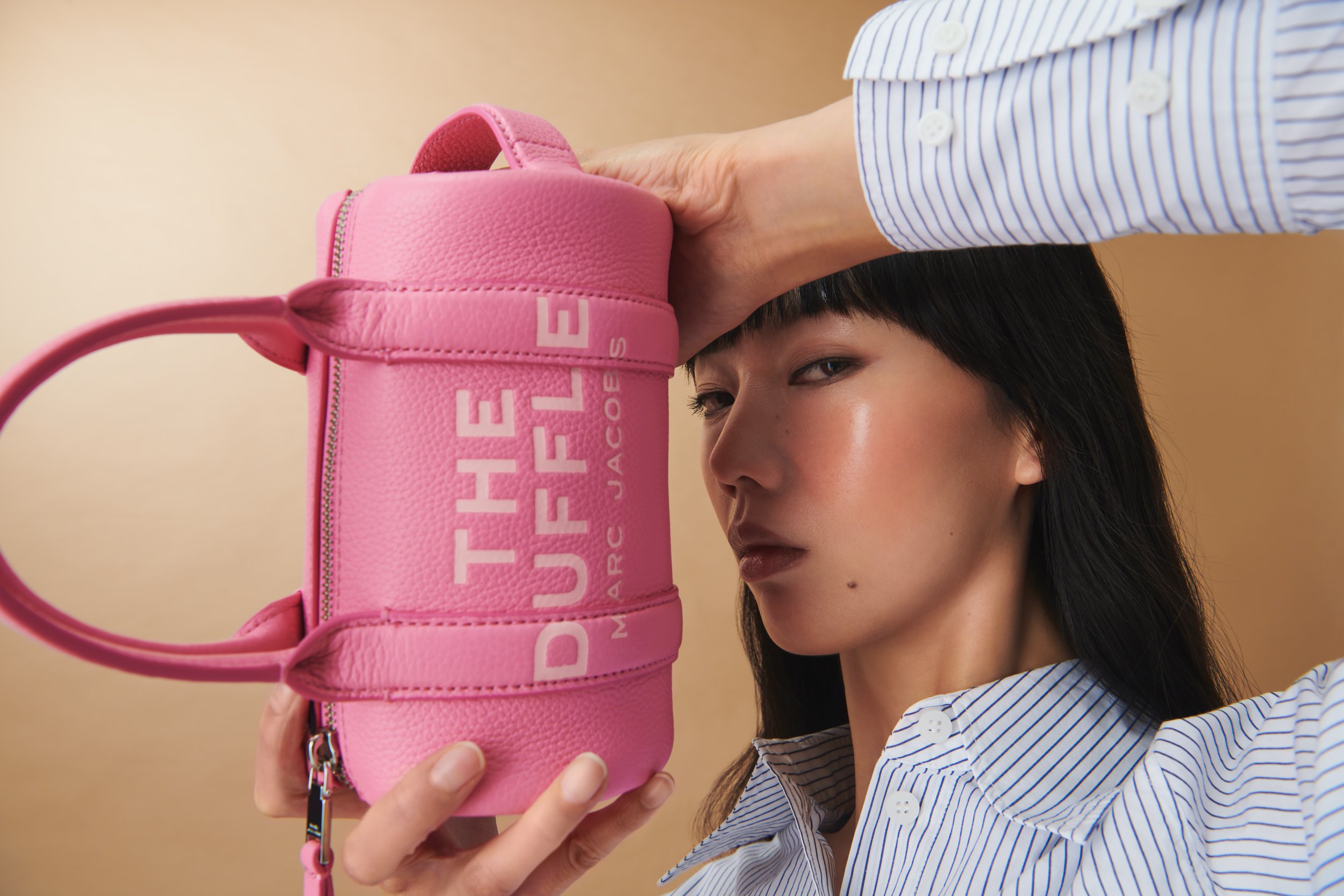 Commercial product photography of Marc Jacobs' pink duffle bag shot by QUEST Production Co., London fashion photographers. Model in a striped shirt holding the bag against a beige background.