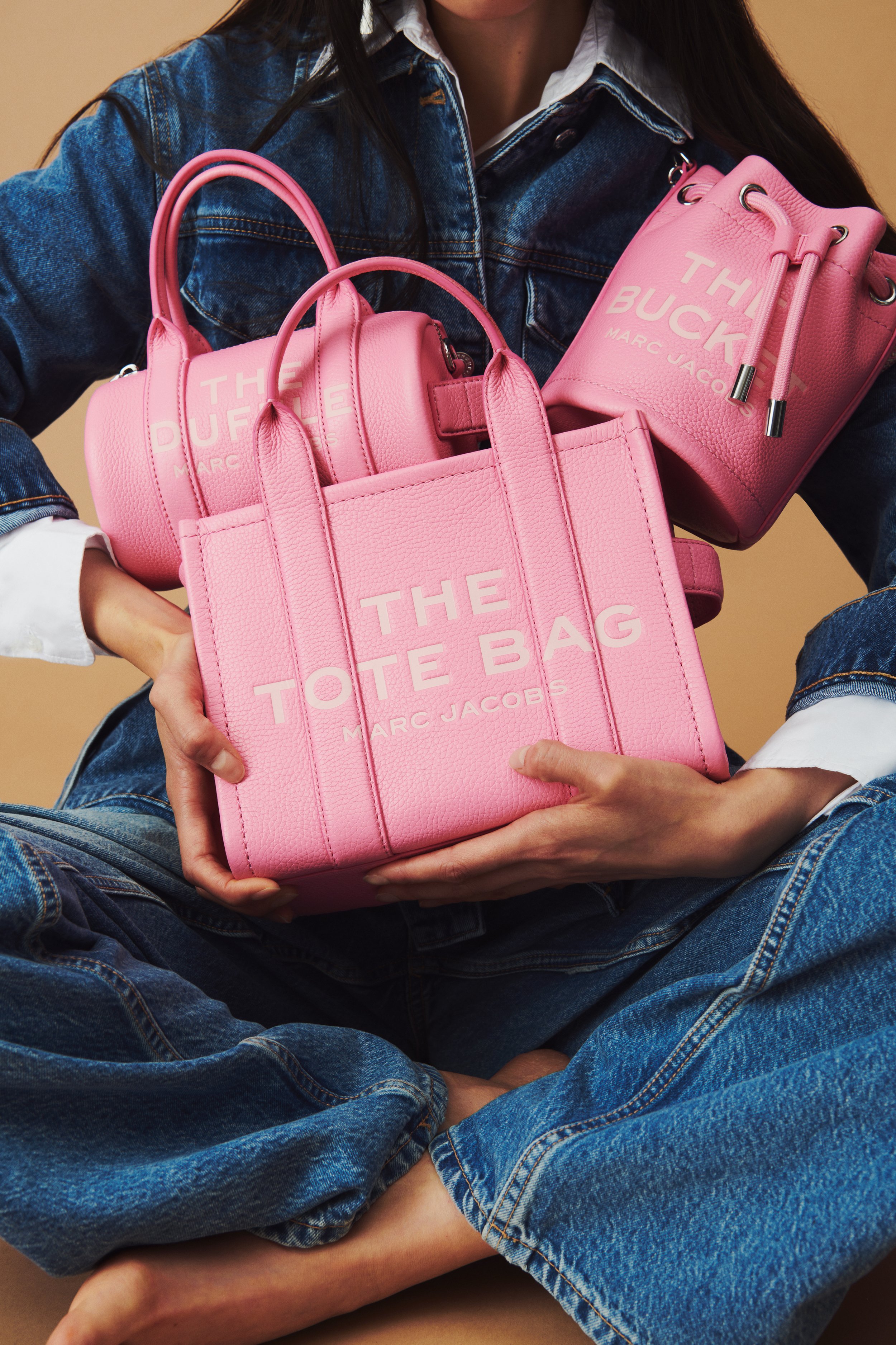 Commercial product photography of Marc Jacobs' pink tote, bucket, and duffle bags shot by QUEST Production Co., London fashion photographers. Model in a denim outfit holding the bags against a beige background.