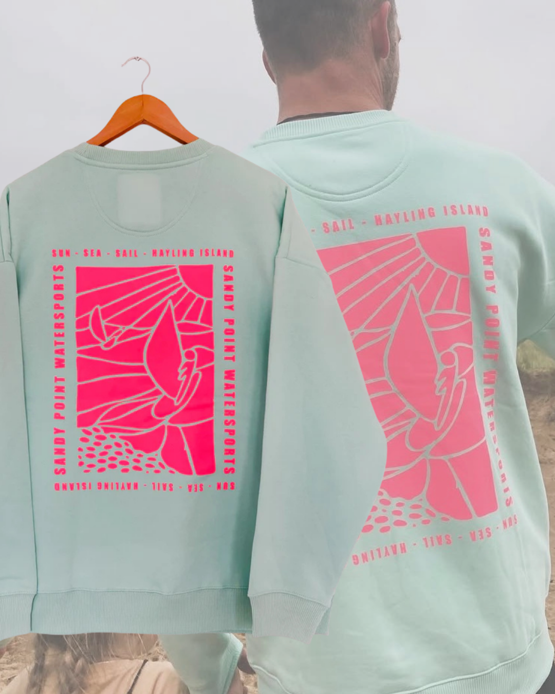 Two people wearing matching light green sweatshirts with a pink graphic and text related to coastal activities on the back, standing outdoors.