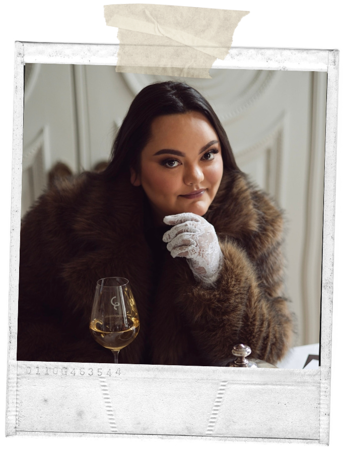 A young woman with dark hair, wearing a brown fur coat and white lace gloves, sitting at a table with a glass of white wine, looking confidently at the camera.