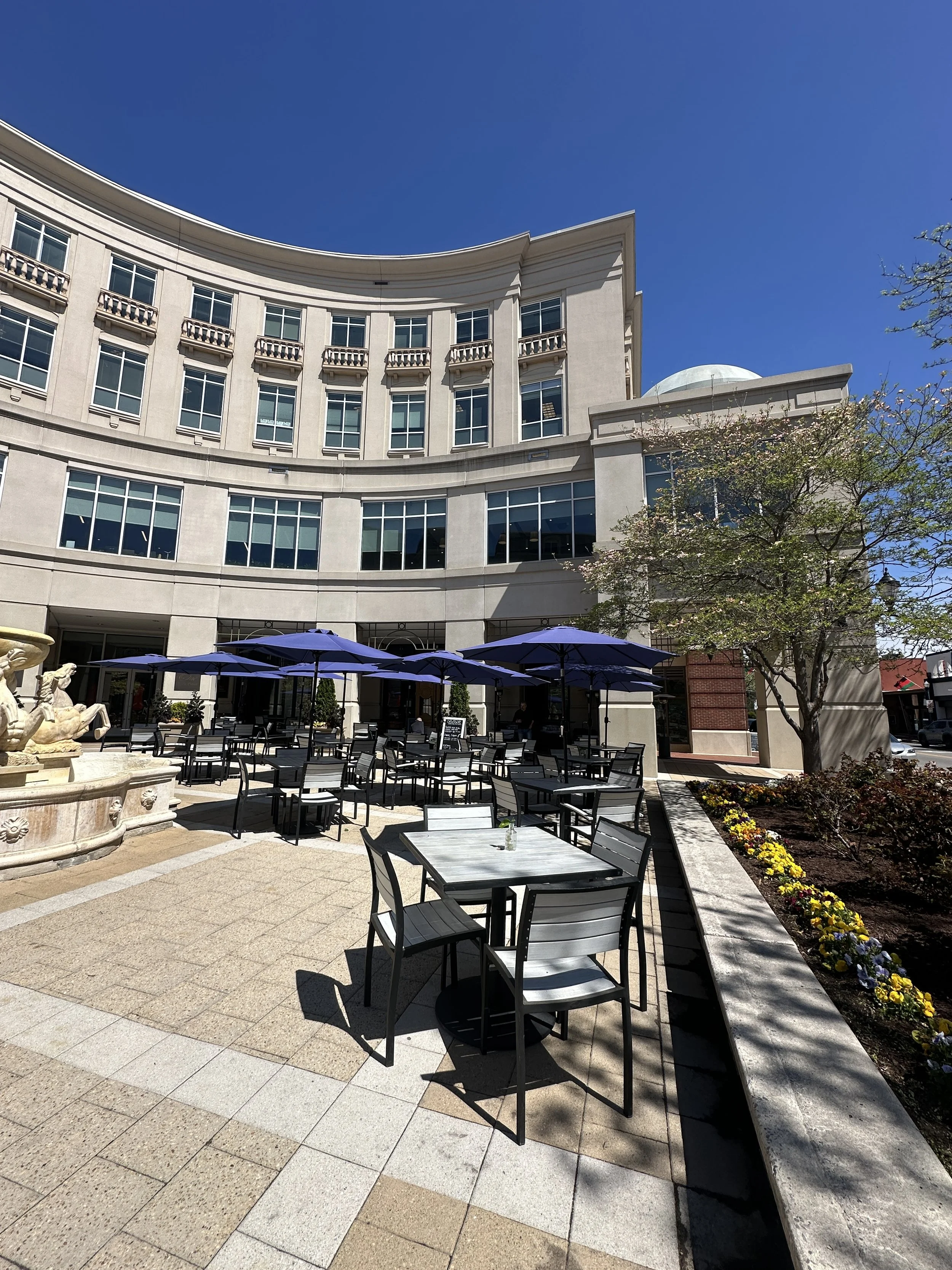 Outdoor seating area with black tables and chairs, blue umbrellas, a fountain with statues, a sidewalk, a flowering tree, and a large beige building with multiple windows against a blue sky.