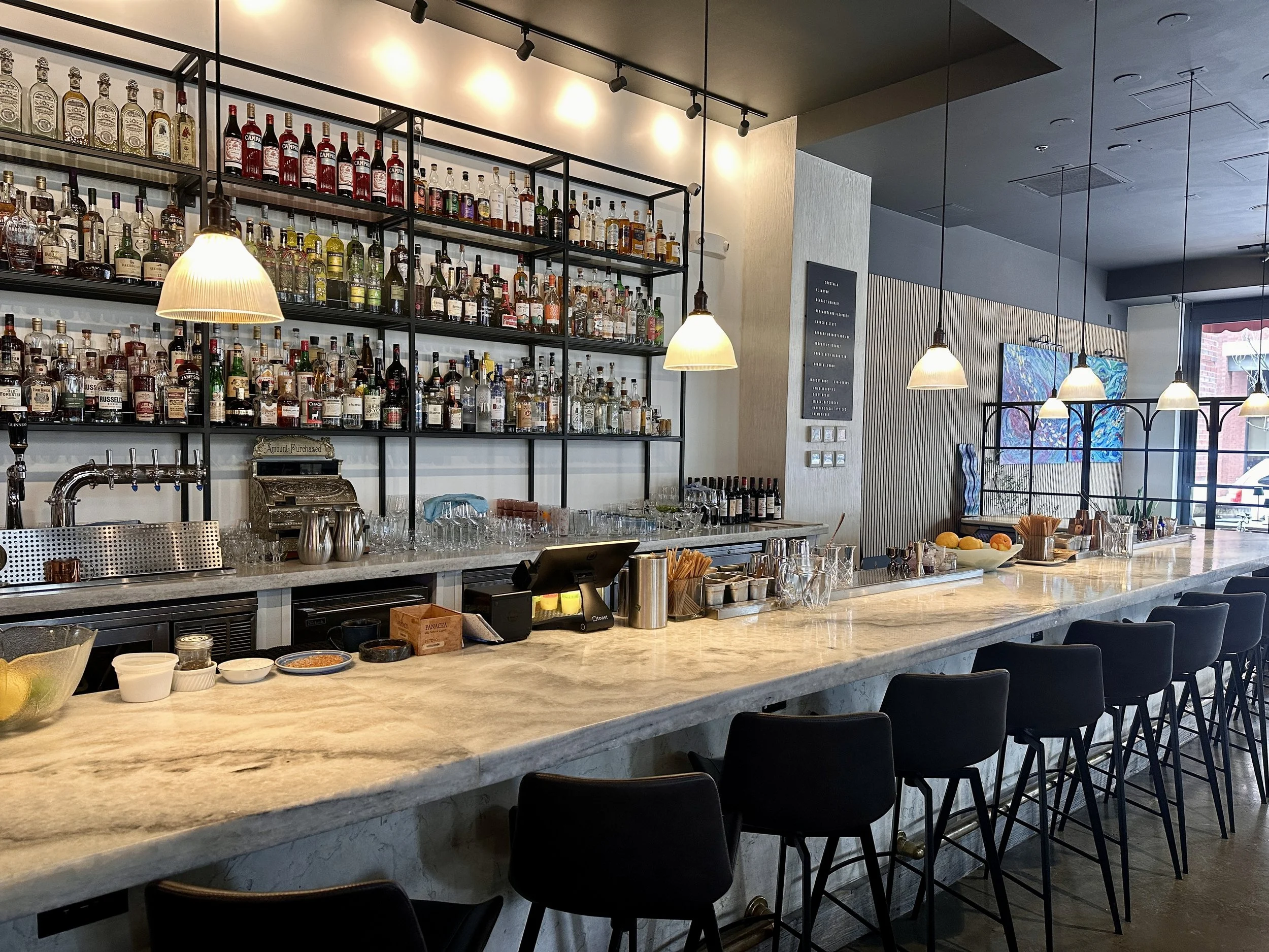 A bar counter in a restaurant or cafe with shelves of various bottles of alcohol behind it. There are three pendant lights hanging above the bar, illuminating the area. The counter has glasses, bowls, and other bar equipment, with a few chairs lined 