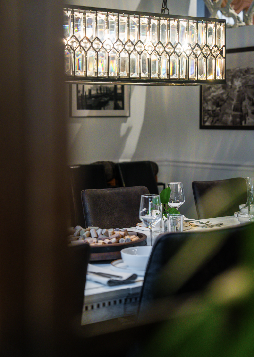 A dining room with a chandelier hanging above a table set with wine glasses, plates, and silverware, with framed photographs on the white walls.