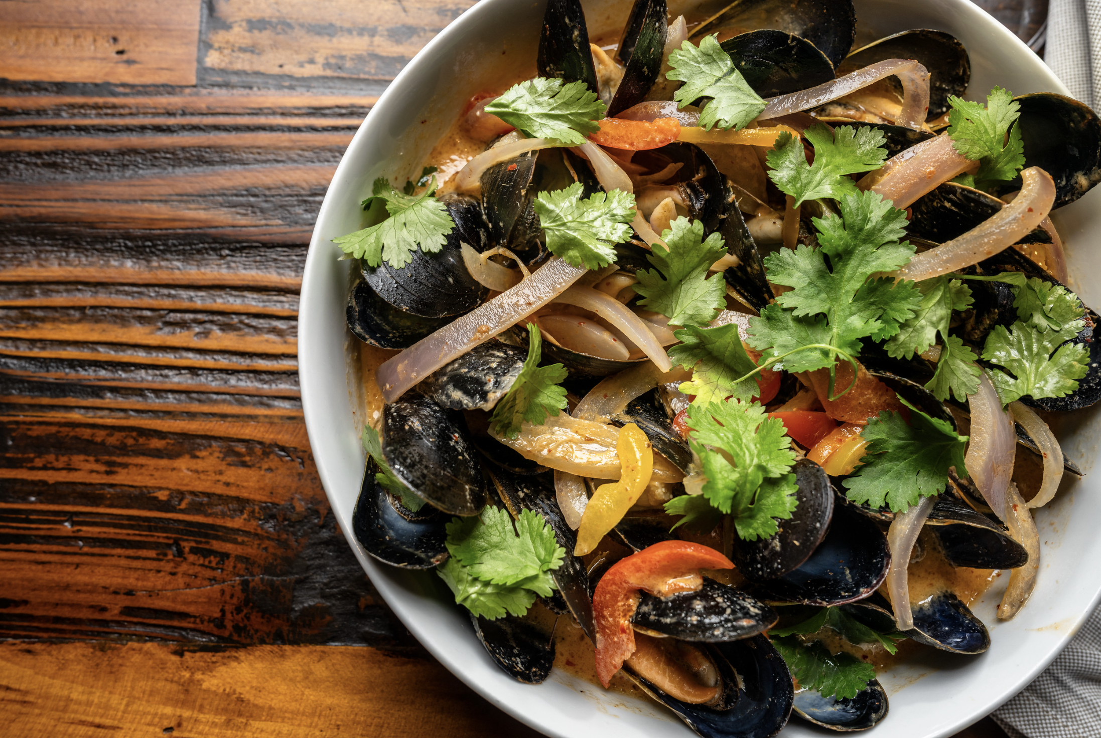 A bowl of mussels with sliced onions, red and yellow bell peppers, garnished with fresh cilantro, on a wooden table.