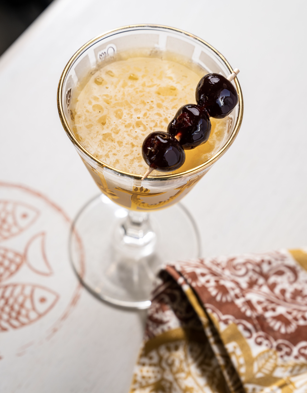 A classic cocktail garnished with three dark cherries on a skewer, served in a stemmed glass on a white surface with a patterned cloth nearby.