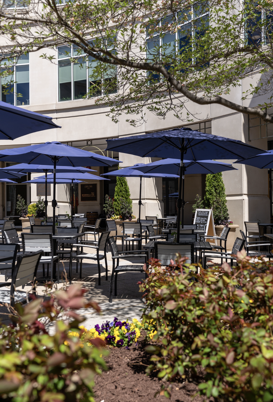 An outdoor patio of a restaurant or cafe with several black tables and chairs, blue umbrellas, potted plants, and colorful flowers, with a modern building in the background.