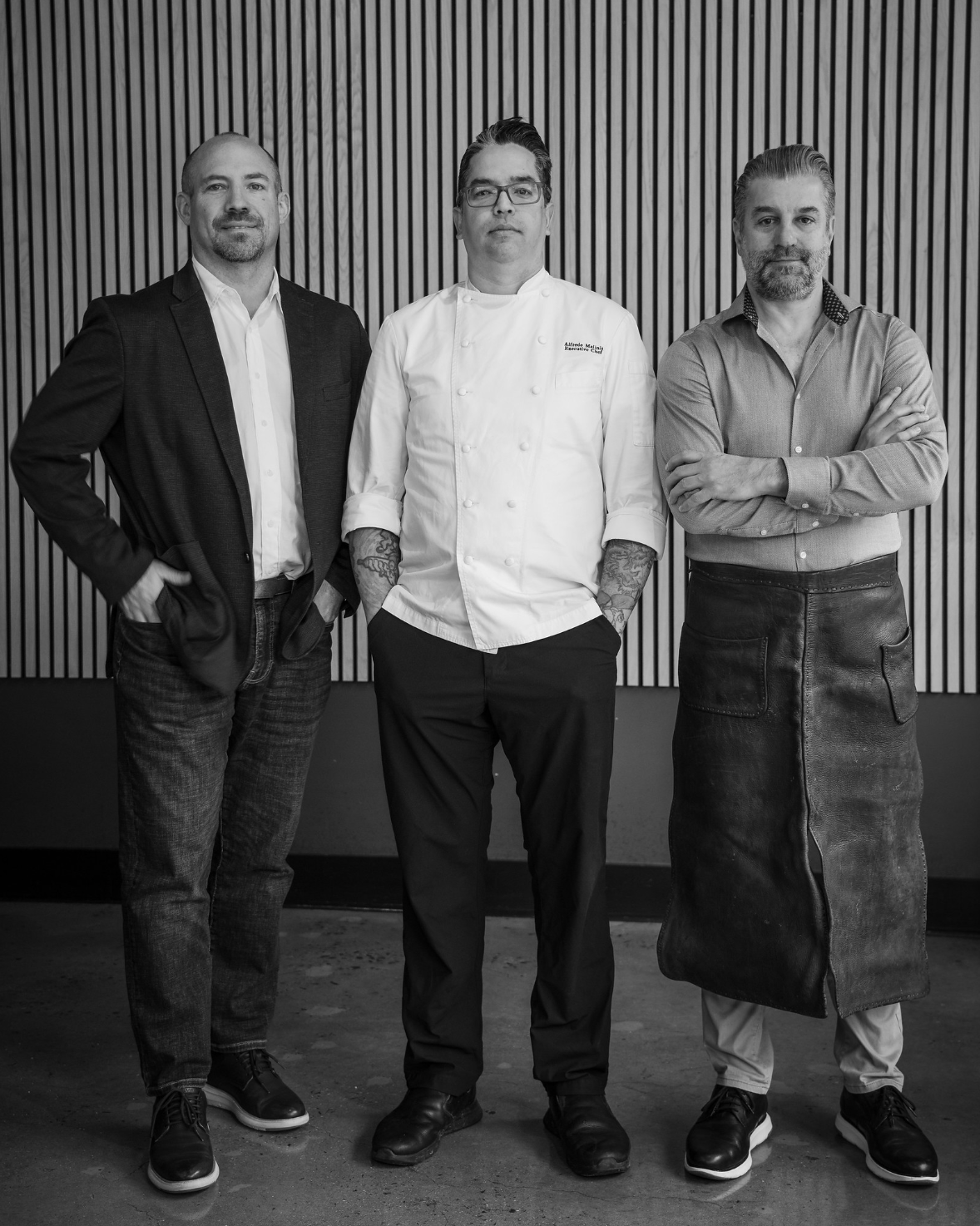 Three men standing side by side in front of a wooden wall, one in a suit, one in a chef's coat, and one in a shirt and apron.