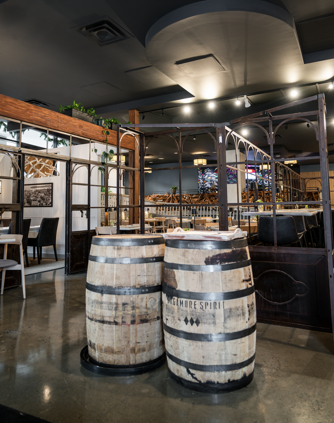 Interior of a restaurant with wooden and metal decor, wooden barrels with 'Orgamoore Spirit' branding, and seating areas divided by metal partitions. There are indoor plants and artwork on the walls.