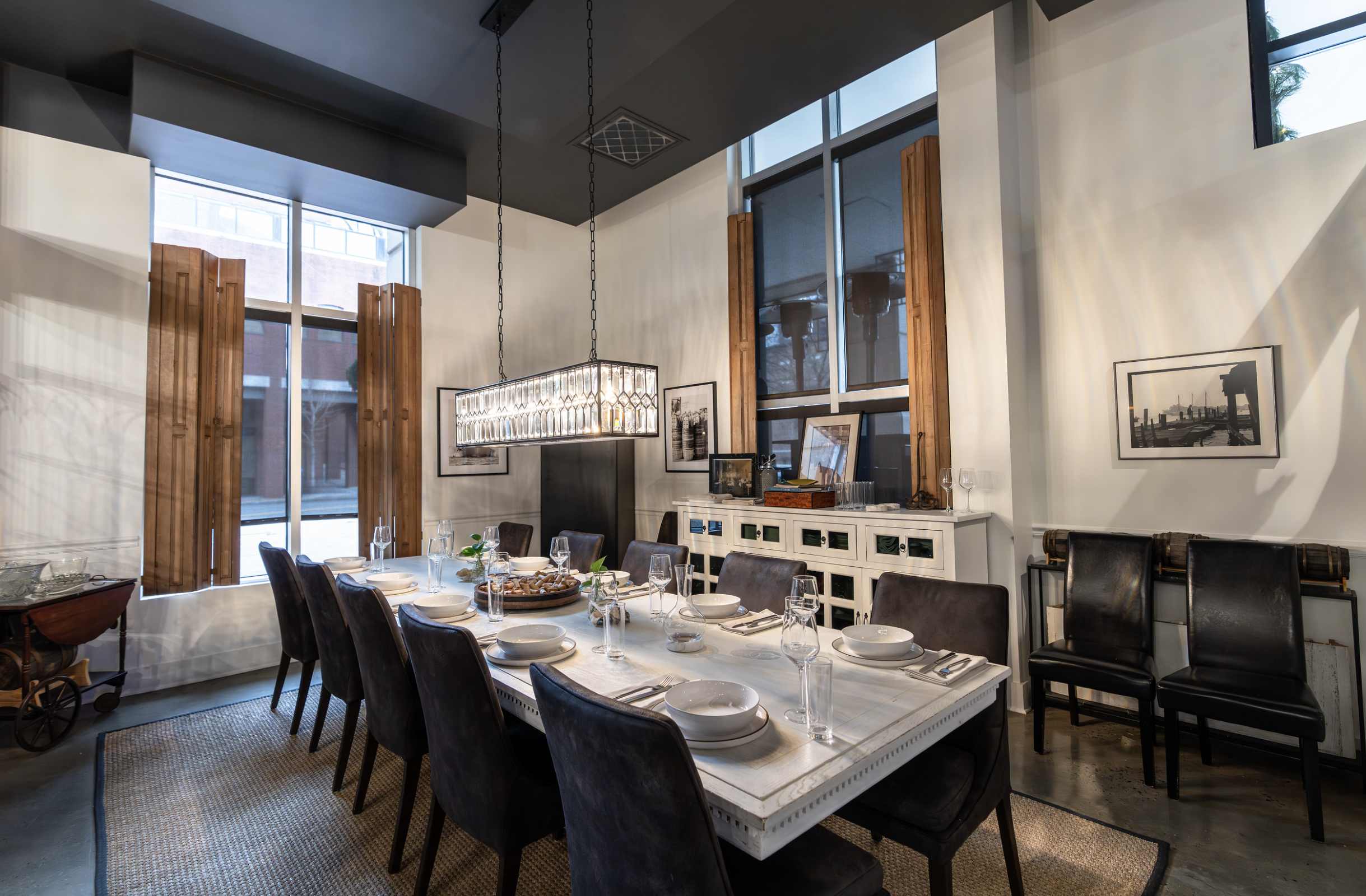 A dining room with a large oval table set with white bowls, glasses, silverware, and a tray of bread. The room has tall windows with wooden shutters, framed art on the walls, and a modern rectangular chandelier hanging above the table.