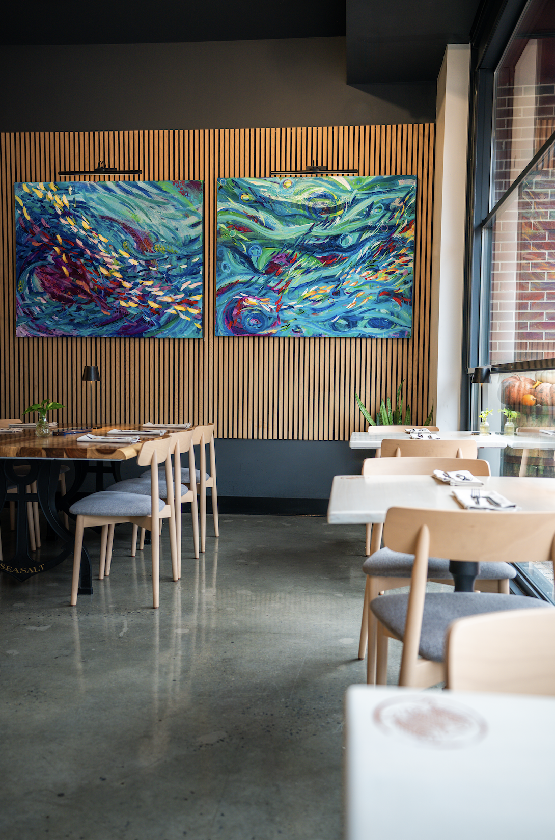 Interior of a modern restaurant with wooden wall paneling, two colorful abstract paintings, and white tables set with napkins and utensils. Large windows allow natural light inside, and there are green plants on the tables.