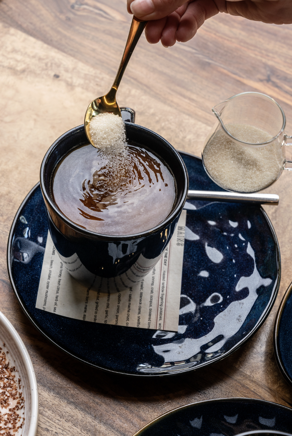 A hand holding a gold spoon adding sugar to a black mug filled with a hot beverage, on a tray with a blue and white abstract patterned plate, a glass pitcher with sugar, a spoon, and part of a dessert dish visible.